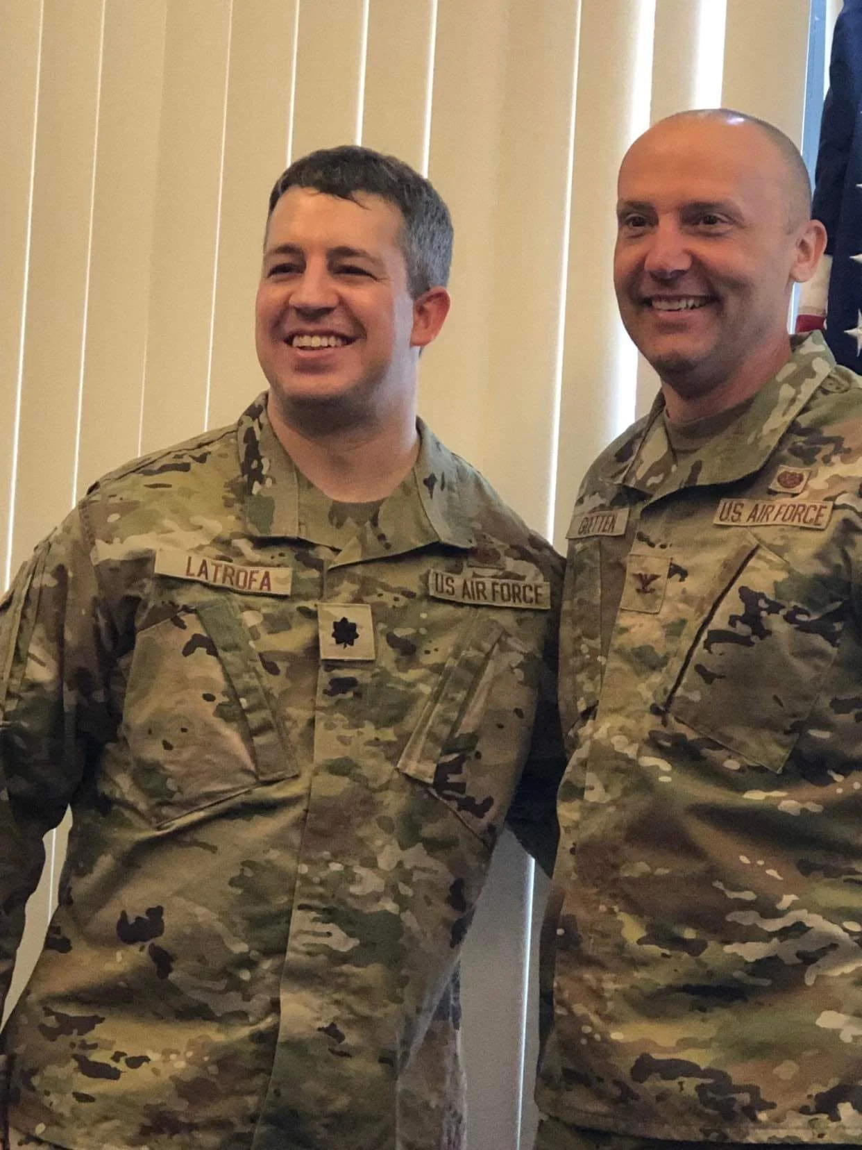Two men in U.S. Air Force uniforms standing side by side smiling at an indoor event, with vertical blinds in the background.