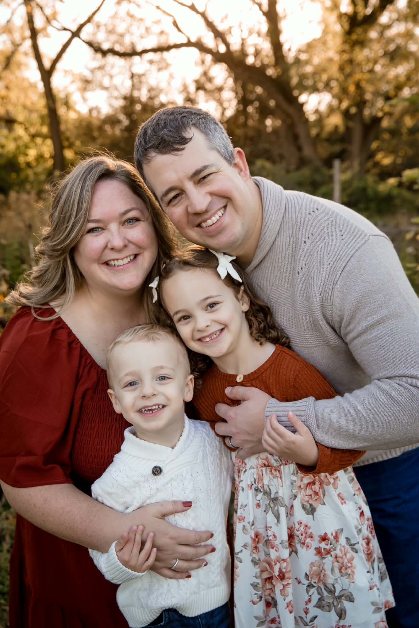 A smiling family of six people, including two adults and four children, posing outdoors in a park with autumn trees in the background.