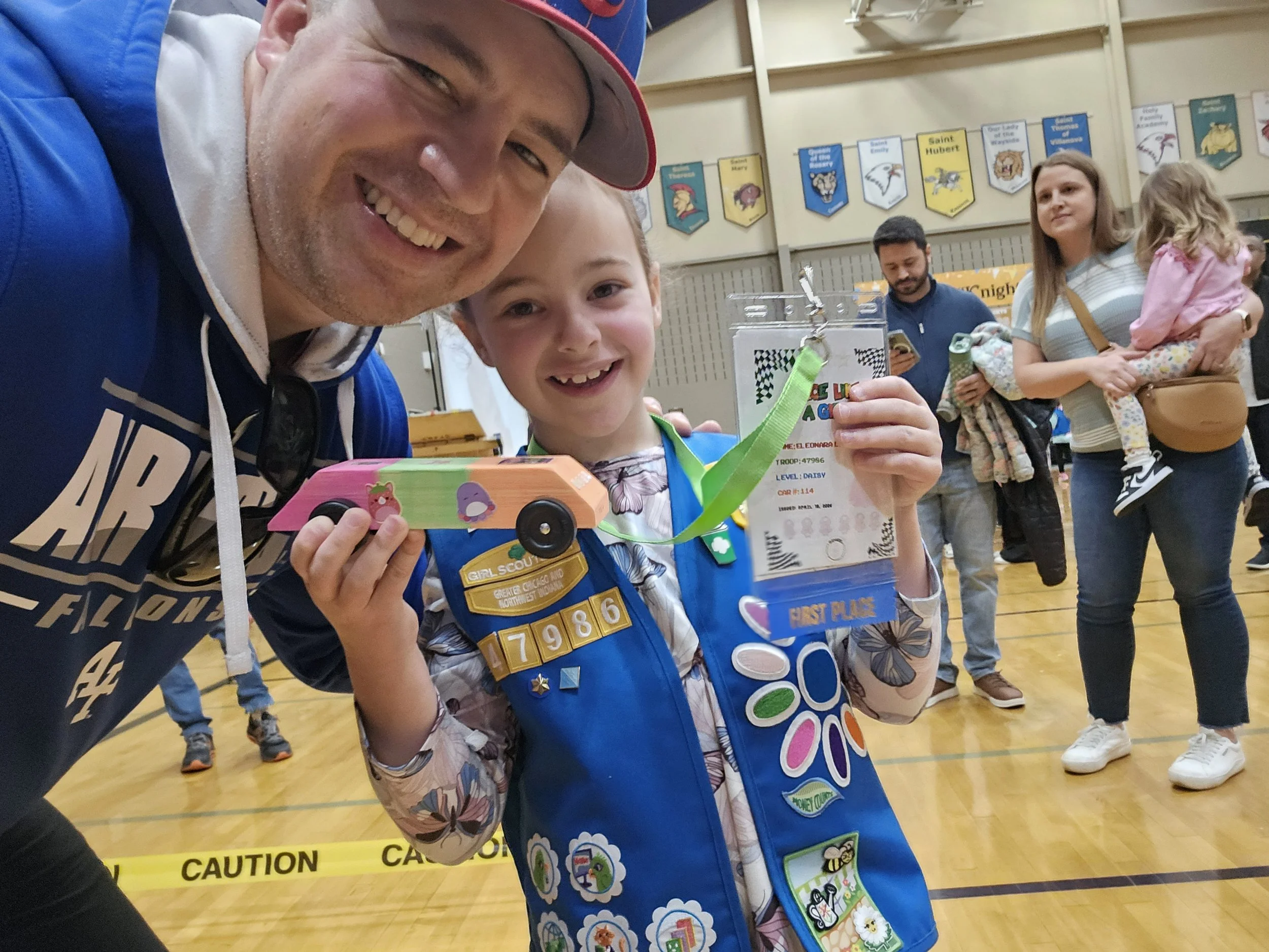A smiling man and boy, possibly a father and son, at a girl scout event in a gymnasium. The boy is holding a small painted wooden vehicle and a trophy ribbon that reads 'First Place.' The boy wears a blue scout vest with various badges, and a girl scout sash with patches. In the background, there are people, and banners with various troop names are hanging on the wall.
