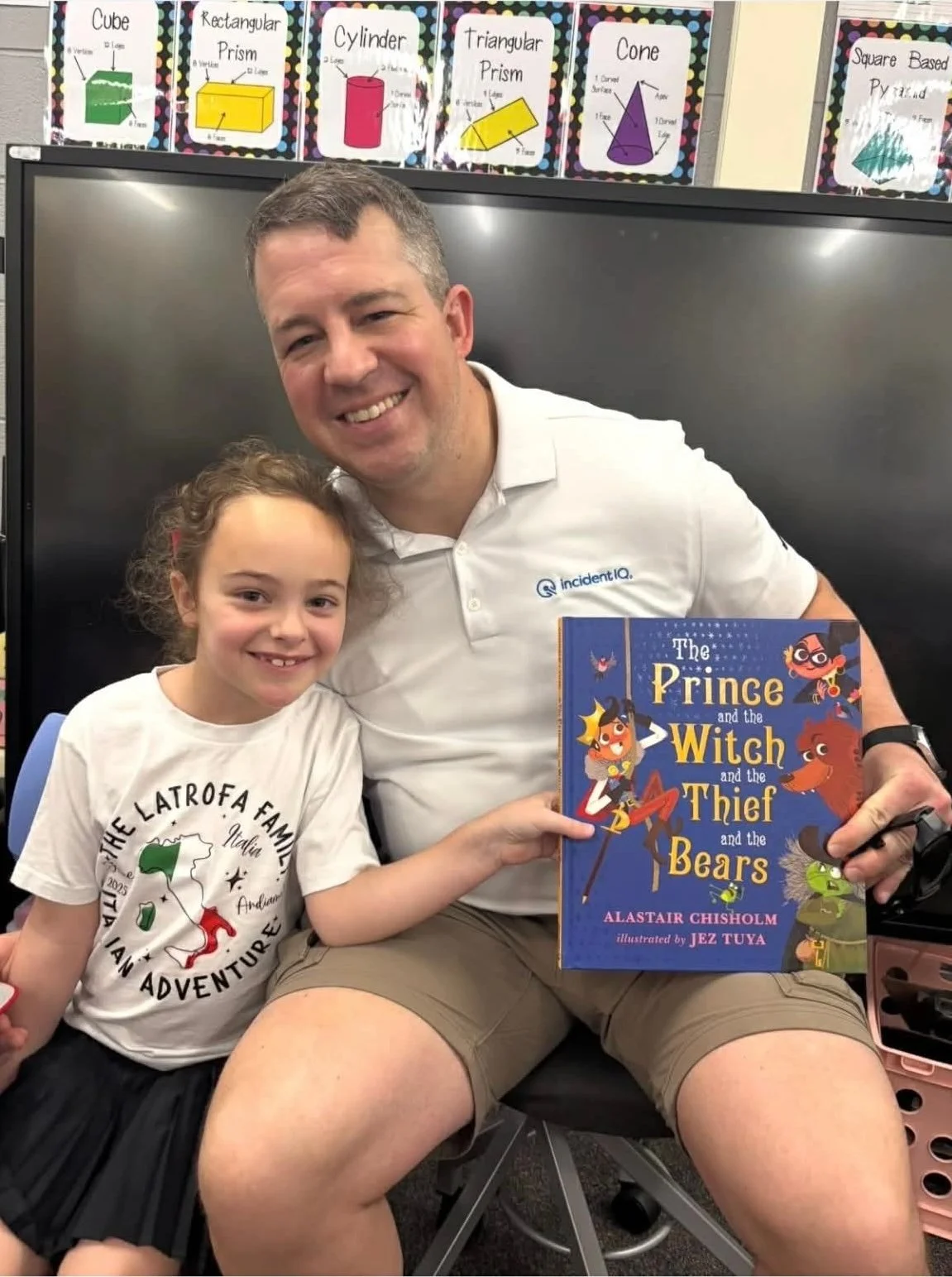 A man and a young girl sitting together, smiling at the camera. The man is holding a children's book titled "The Prince and the Witch and the Thief and the Bears." The girl is wearing a white T-shirt with a map of Italy and the words "The Latrofa Family Italian Adventure." Behind them, there are educational posters about 3D geometric shapes on the wall.