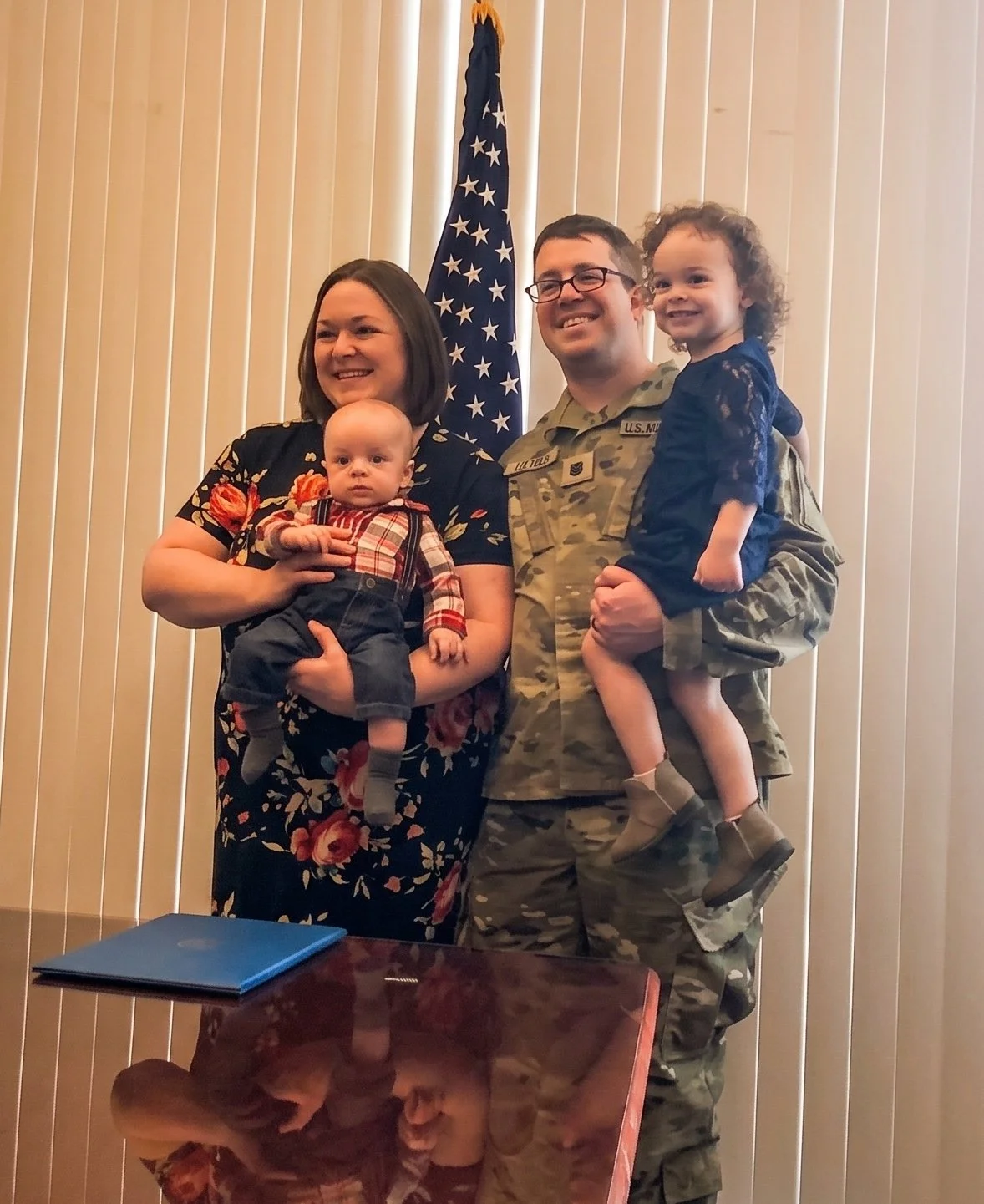 Family photo with candidate in military uniform, standing in front of an American flag and vertical blinds.