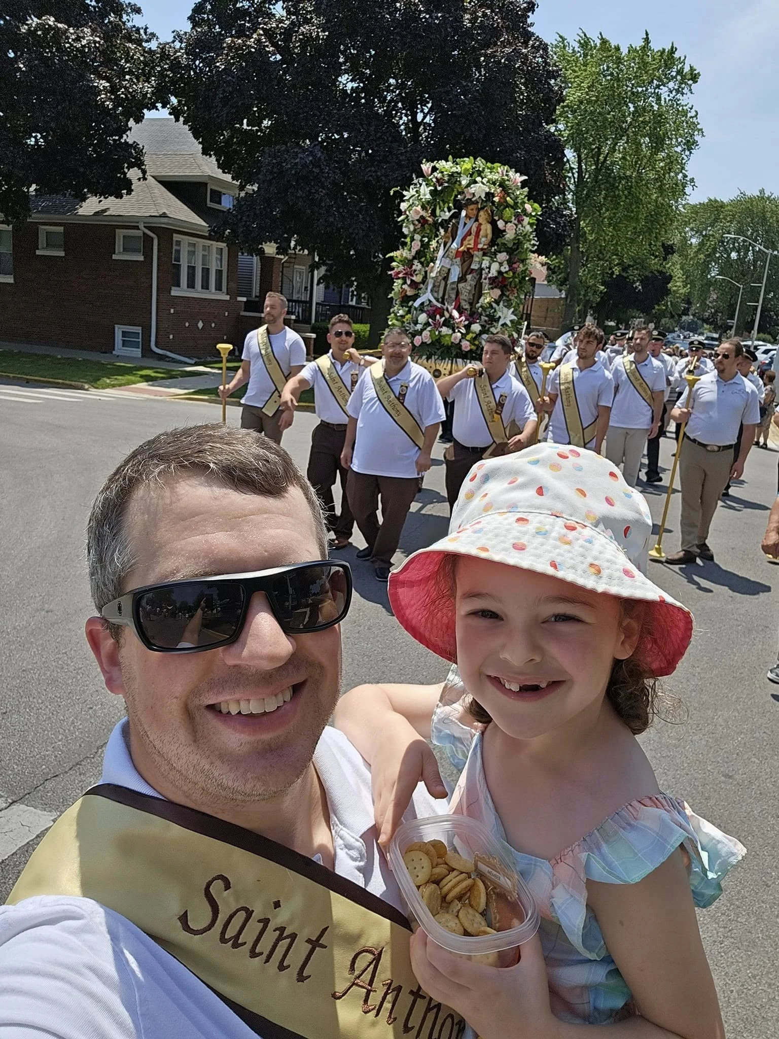 A man and a young girl taking a selfie during a parade. The man is smiling and wearing sunglasses, while the girl is holding a container of cookies. In the background, there are people marching in the parade behind a float with religious imagery, flowers, and people dressed in white with sashes.
