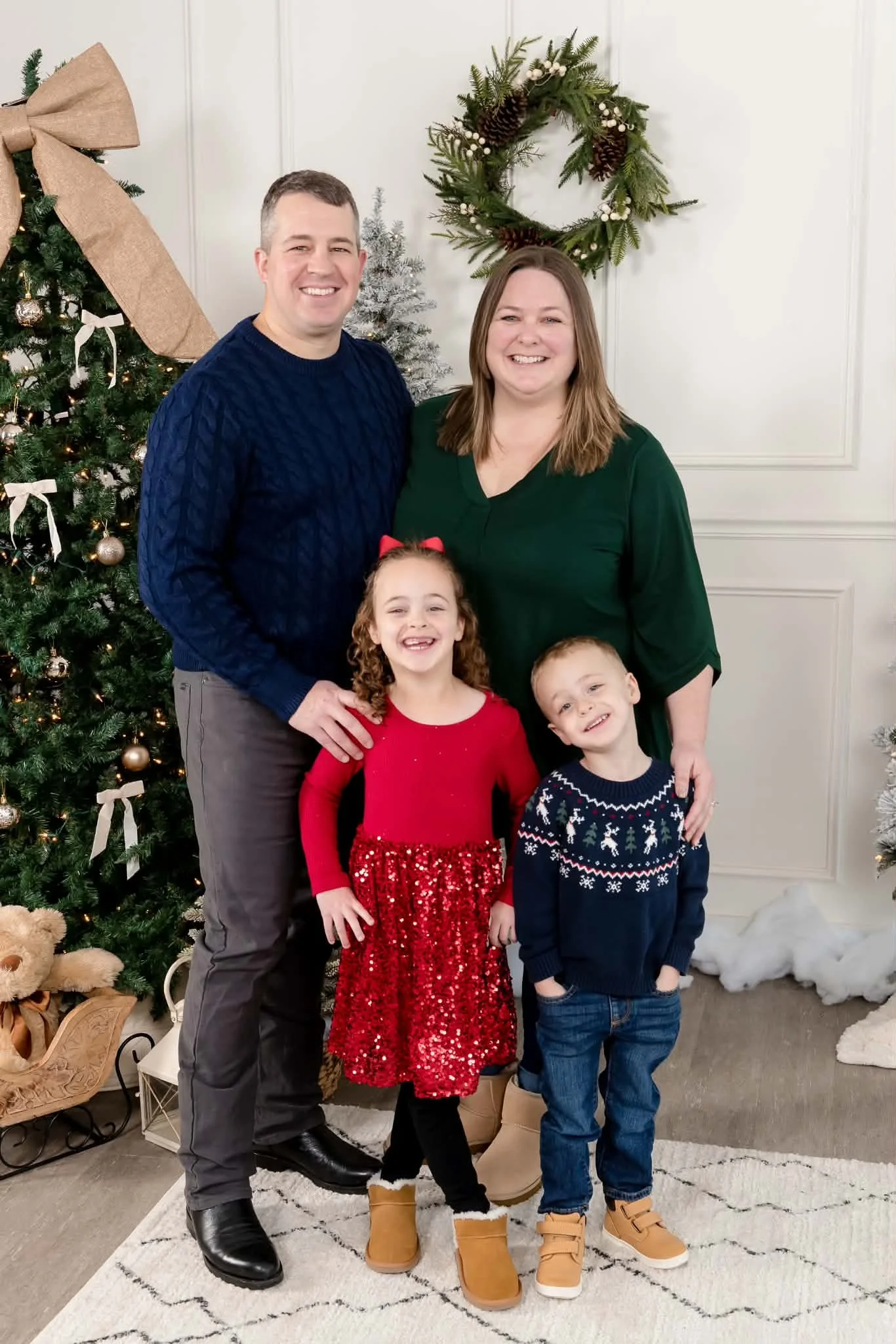 A family of four celebrating Christmas indoors, standing in front of a decorated Christmas tree and a holiday wreath. The family includes a man, a woman, a young girl, and a young boy, all smiling and dressed in festive attire.