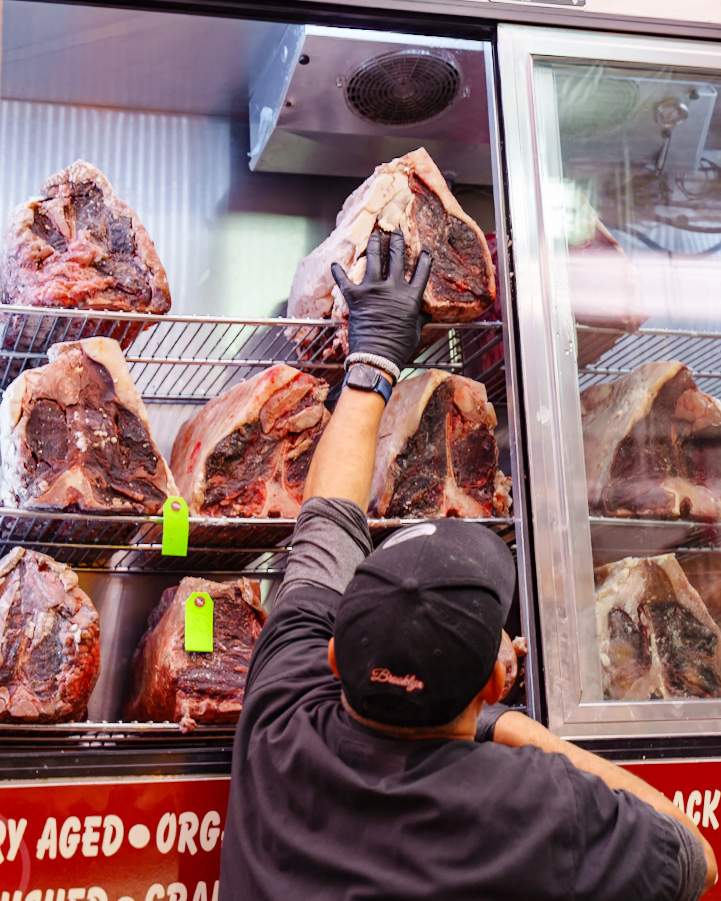 Person in black jacket and black cap handling large cuts of raw aged beef in a meat display case.