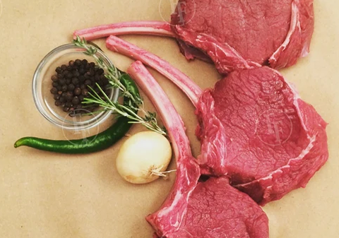 Three raw bison chops, a garlic bulb, black peppercorns in a small bowl, a green chili pepper, and a sprig of rosemary.
