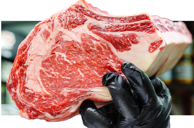 Person wearing black gloves holding a large marbled raw beef steak in a butcher shop.