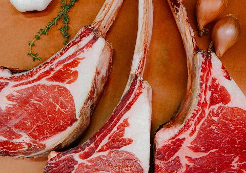 Three raw beef short ribs with marbling, onions, and garlic cloves in the background.
