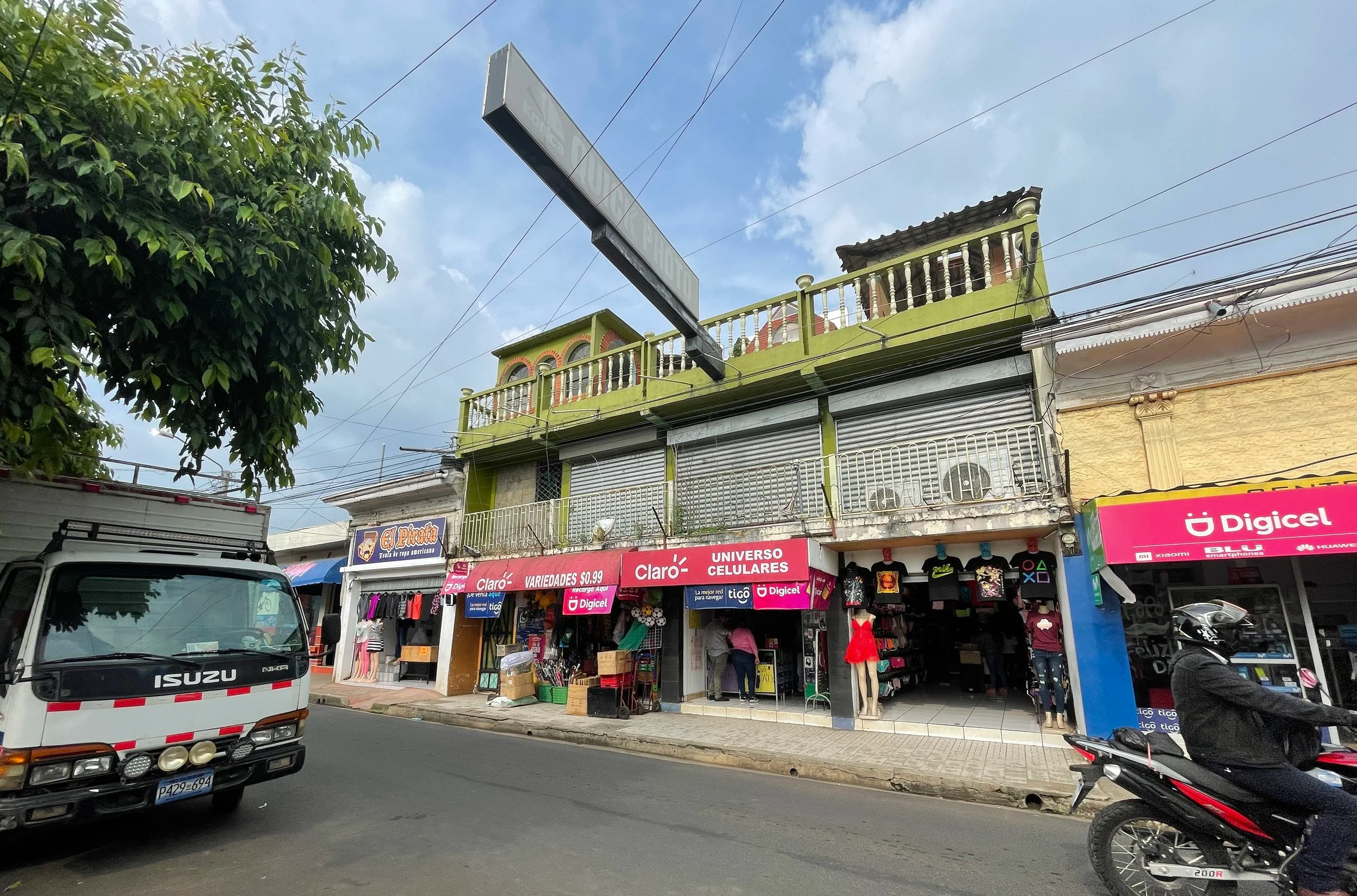 Street scene with small shops selling clothing and accessories, vehicles parked along the sidewalk, including a white Isuzu truck and a bicycle, with a green building featuring a balcony on the upper floor and a clear blue sky overhead.