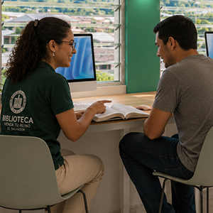 A woman and a man sitting at a desk having a conversation in a library or study area, with computers and large windows in the background.