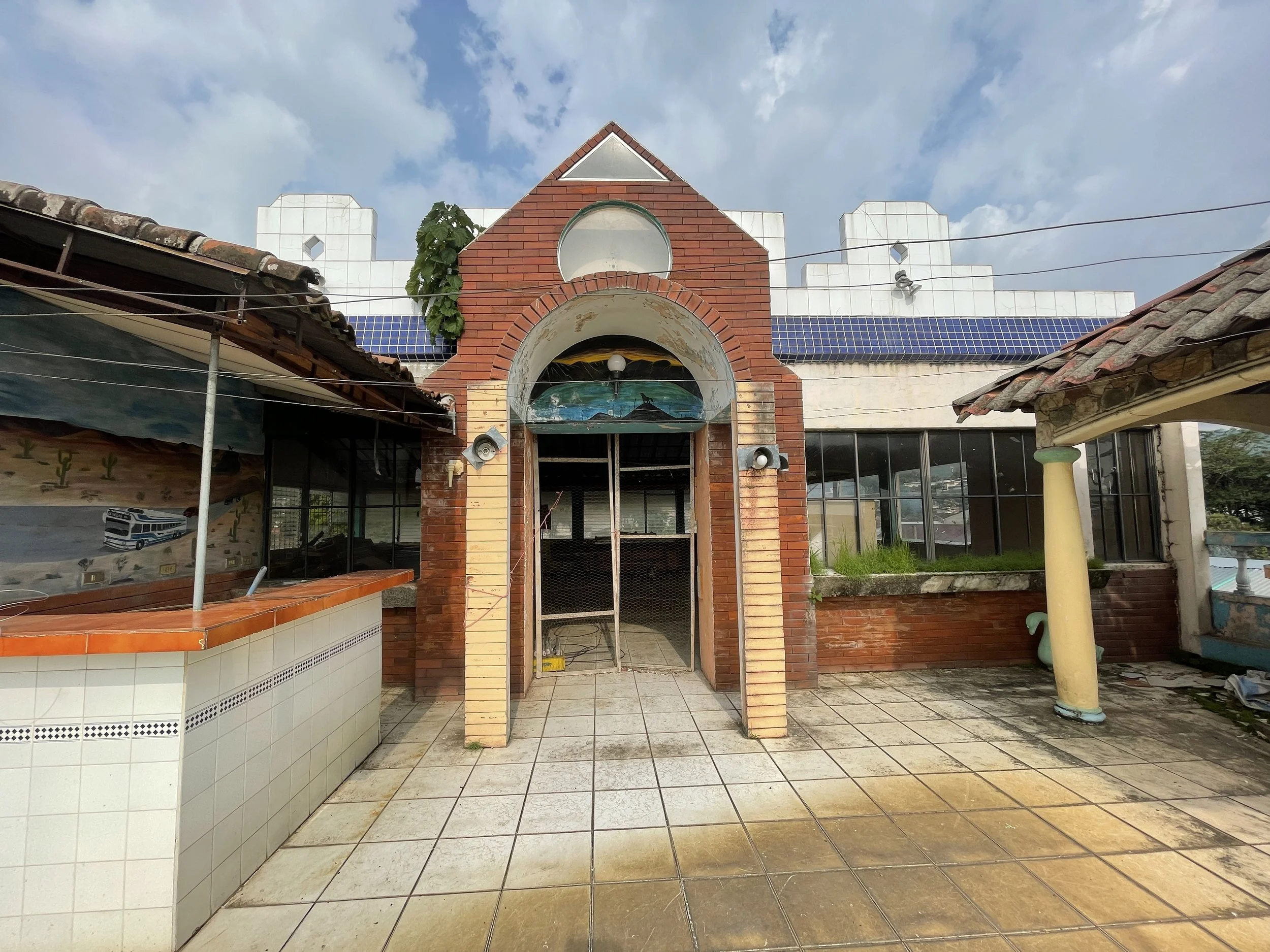 A building under renovation with a brick archway at the entrance, surrounded by tiled flooring, with some roofing tiles and construction materials visible. The sky is partly cloudy.