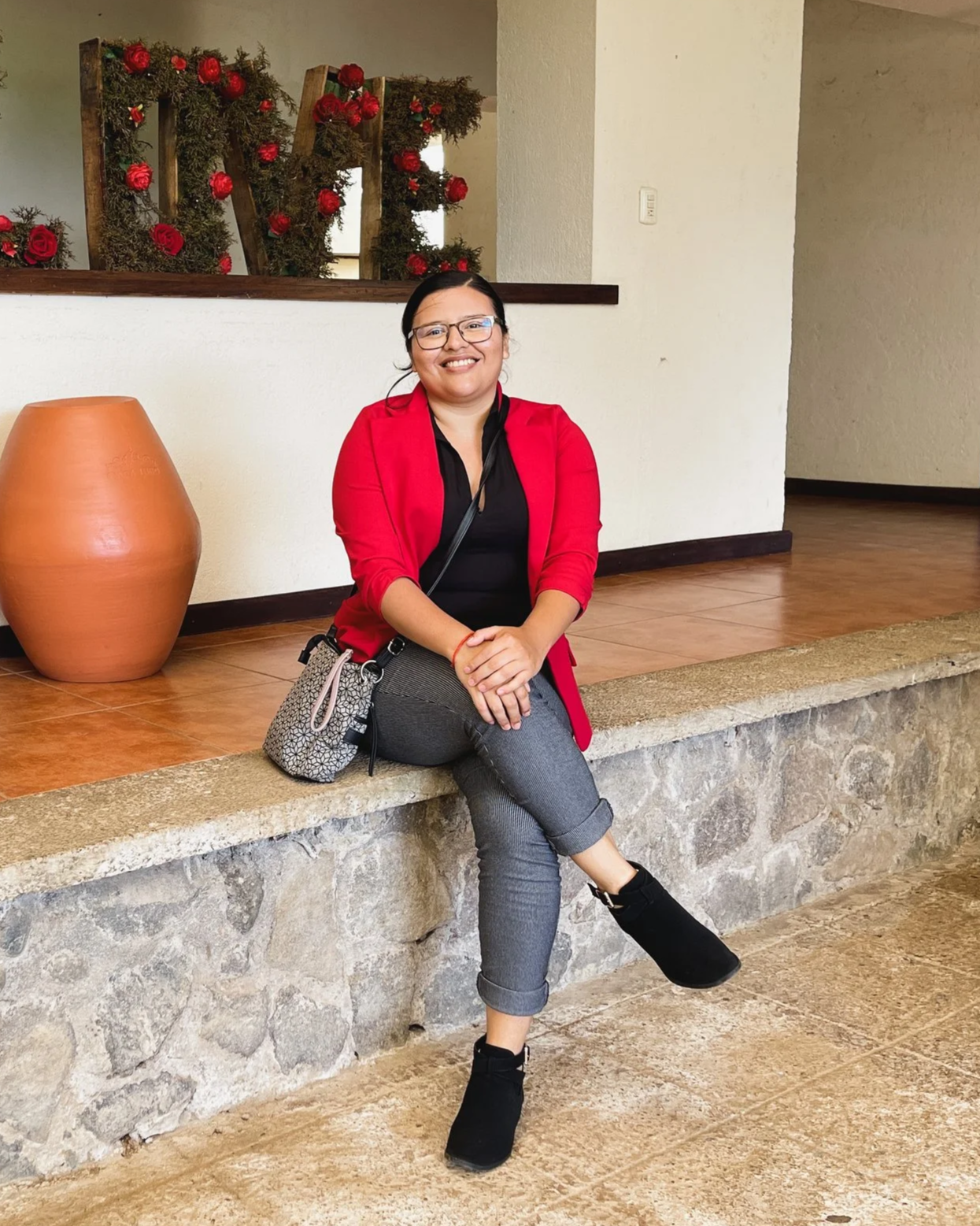 A woman sitting on a stone ledge in an indoor setting, smiling, wearing glasses, a red blazer, black top, gray pants, and black shoes, with a handbag beside her, behind her decorated with red flowers and greenery.