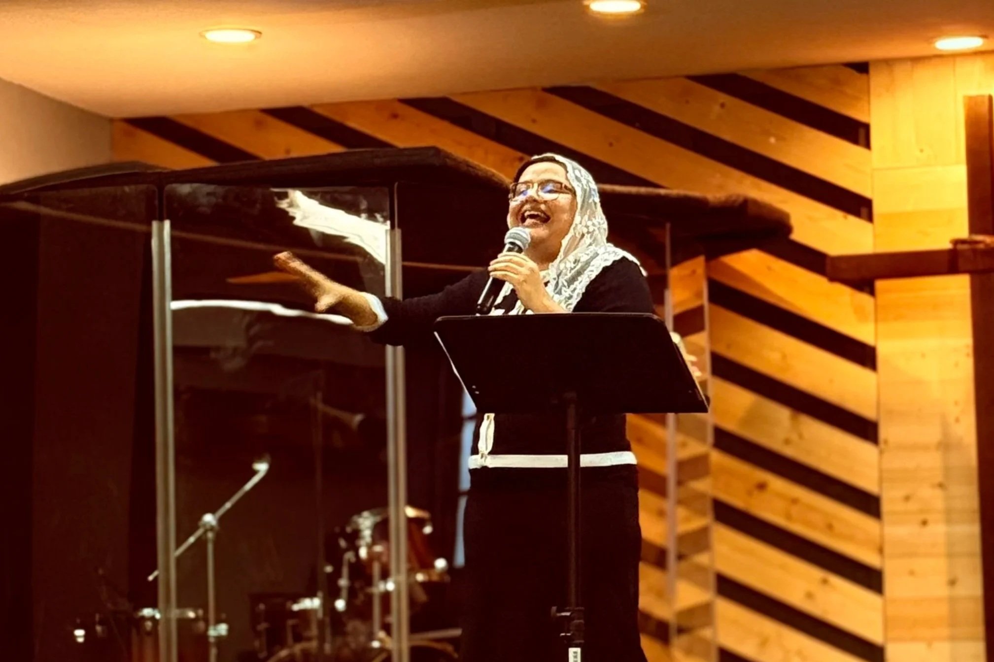 A woman with glasses and a lace headscarf speaking or singing into a microphone, standing in front of a music stand, inside a room with wooden wall decor.