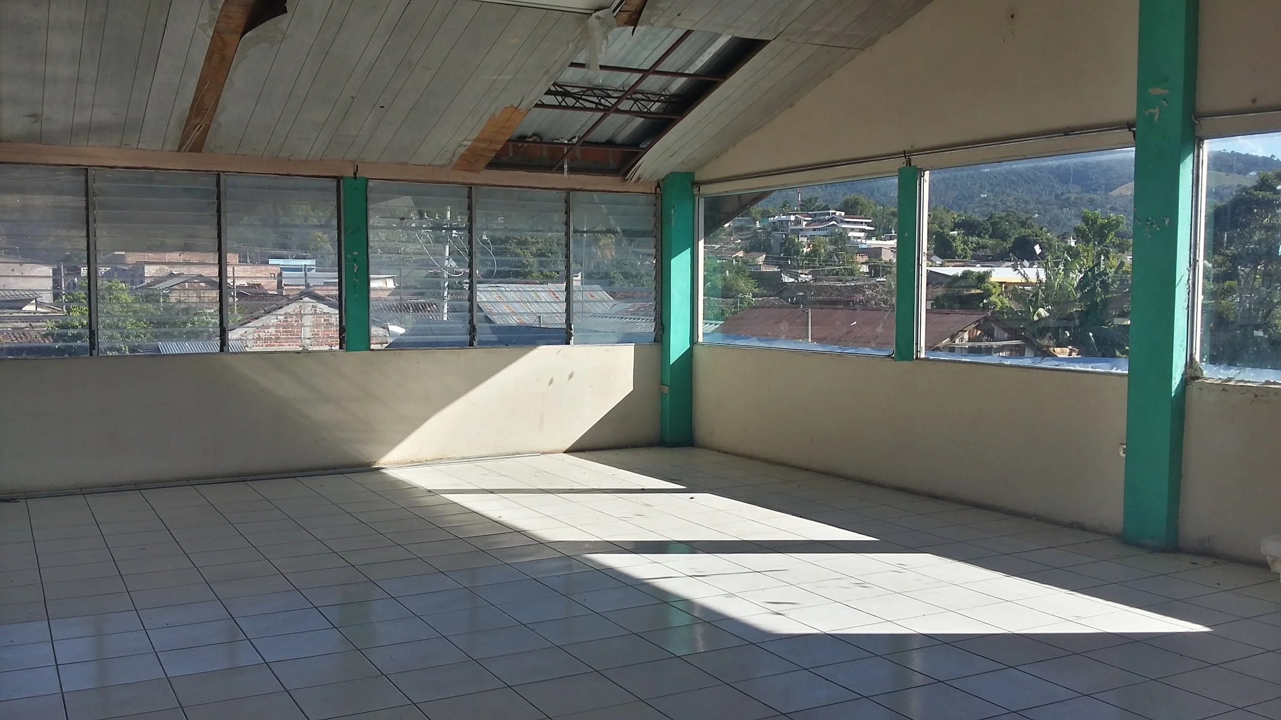 An empty indoor balcony with tiled floor and large glass windows providing a view of houses and greenery outside, with a sloped ceiling and some missing ceiling panels.