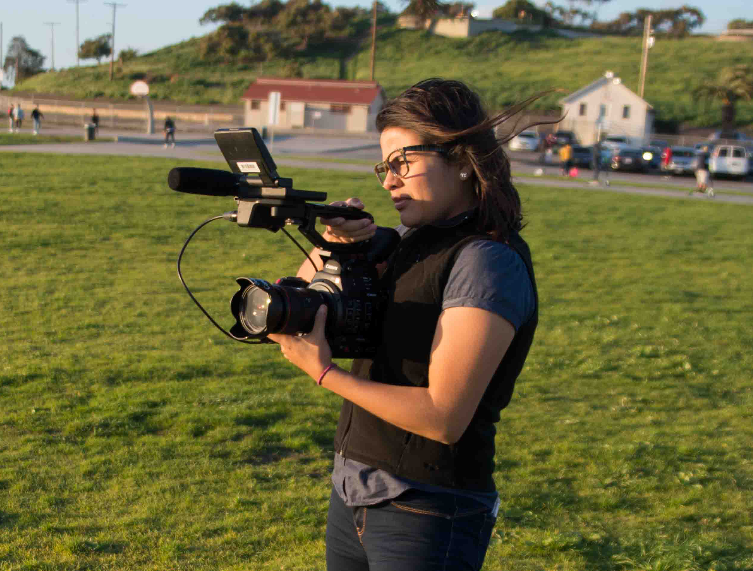A woman with dark hair, glasses, and ear earrings operating a professional video camera outdoors on a grassy field, with people and small buildings in the background during late afternoon or early evening.