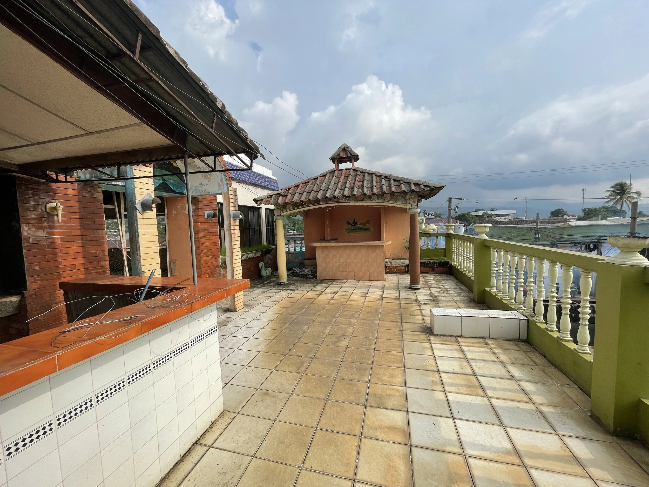 A spacious rooftop terrace with tiled flooring, a small pavilion with a tiled roof, a green railing, and various outdoor decor, under a partly cloudy sky.