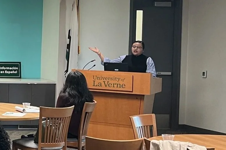 Person giving a presentation or lecture at a podium at the University of La Verne, with audience members seated nearby.