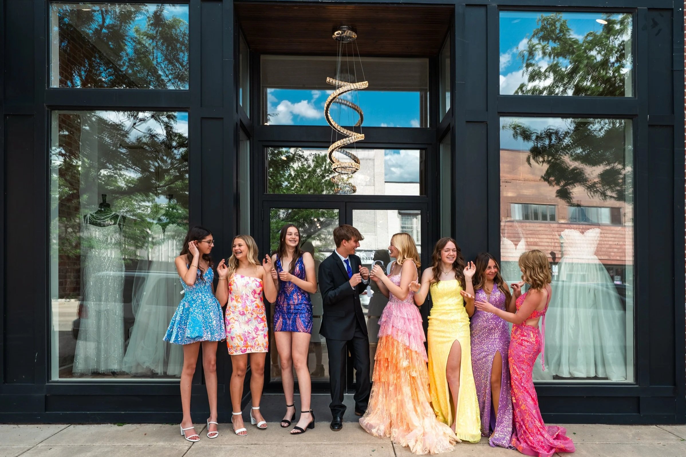 A group of young women and a young man dressed in colorful prom dresses and a suit, standing together outside a modern building with large glass windows. They appear to be celebrating or socializing during a special event, possibly prom or a formal gathering.