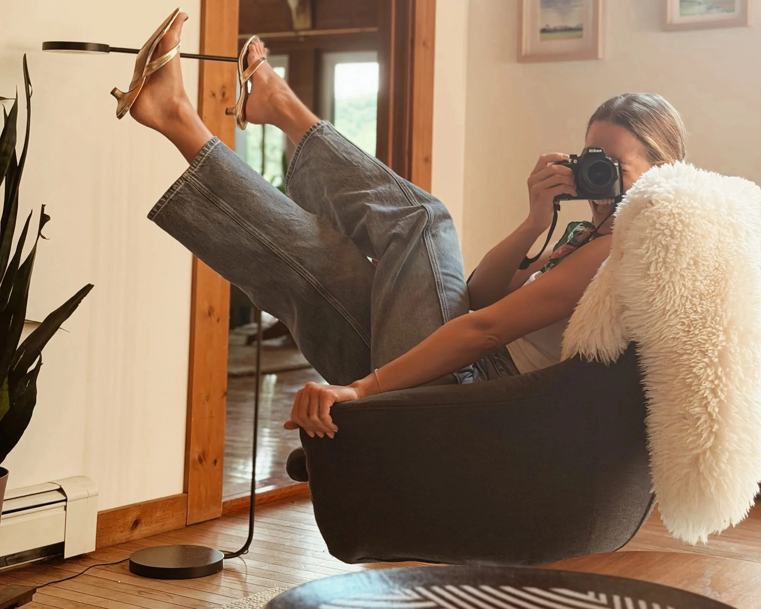 Person taking a photo in a mirror with their phone, legs up on a table, holding a pair of high-heeled shoes, in a room with wooden floors and a wooden door.