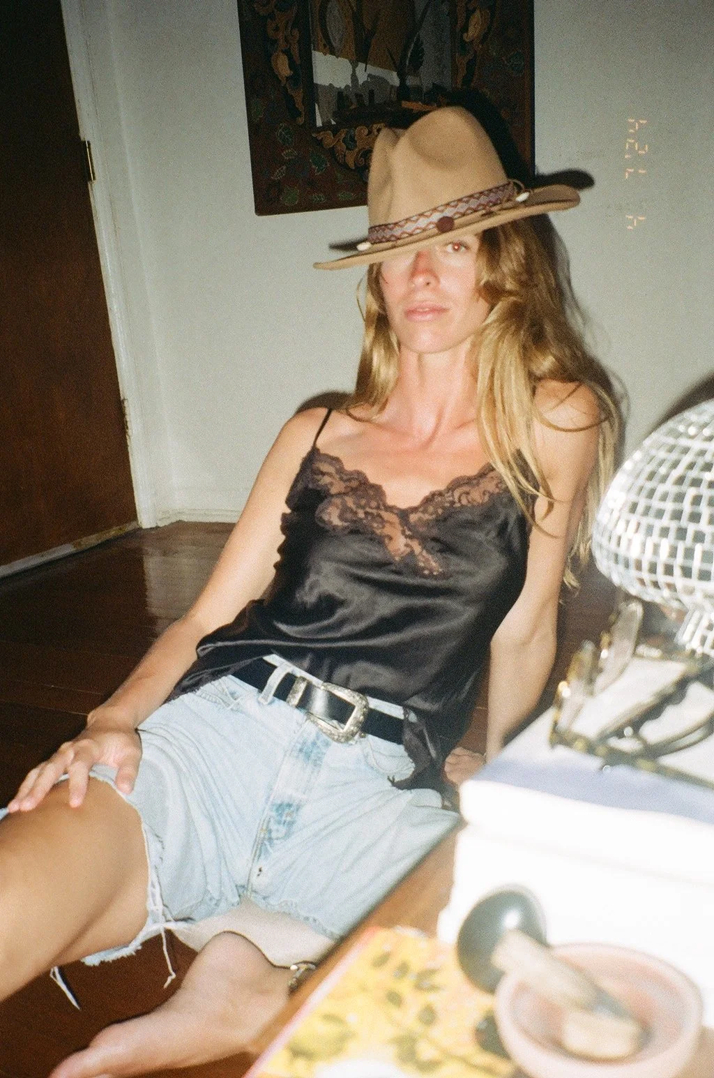 A woman with long blonde hair wearing a tan fedora hat, a black satin camisole with lace trim, and distressed denim shorts, sitting in a room with white walls, wooden floors, and a colorful framed artwork behind her. There are some objects in the foreground, including a silver desk fan and a magazine.