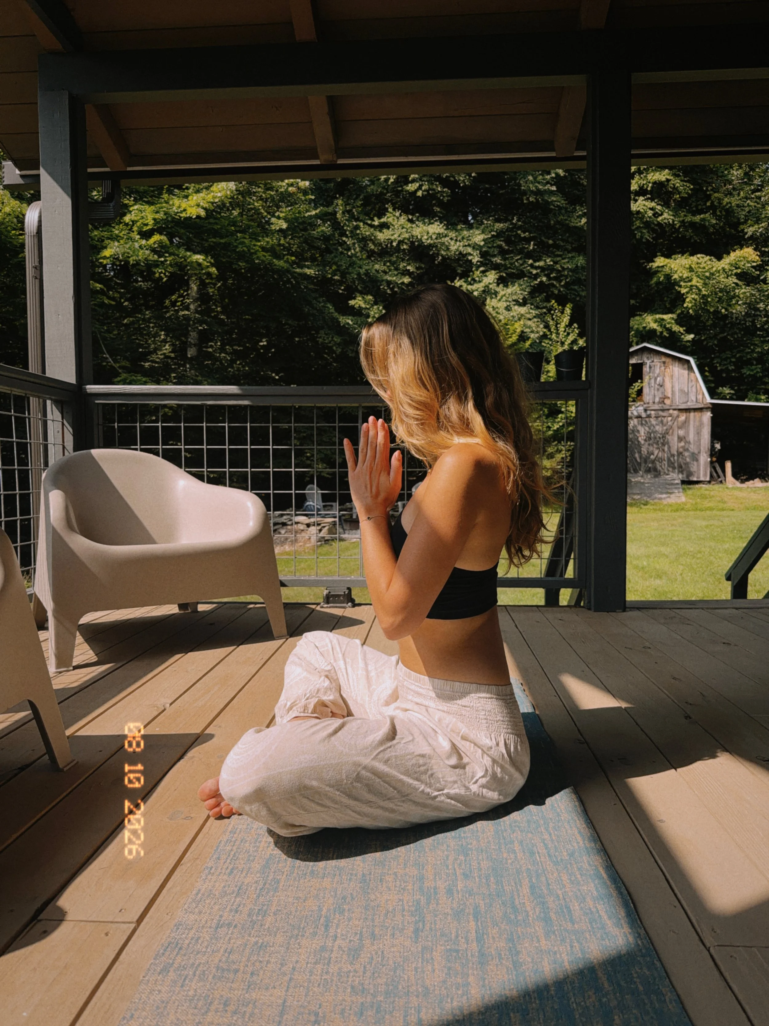 A woman with long hair practicing yoga or meditation outdoors on a wooden deck, sitting cross-legged on a blue and beige rug, with her hands pressed together in a prayer position. She is facing away from the camera, with a background of green trees and a small wooden shed.