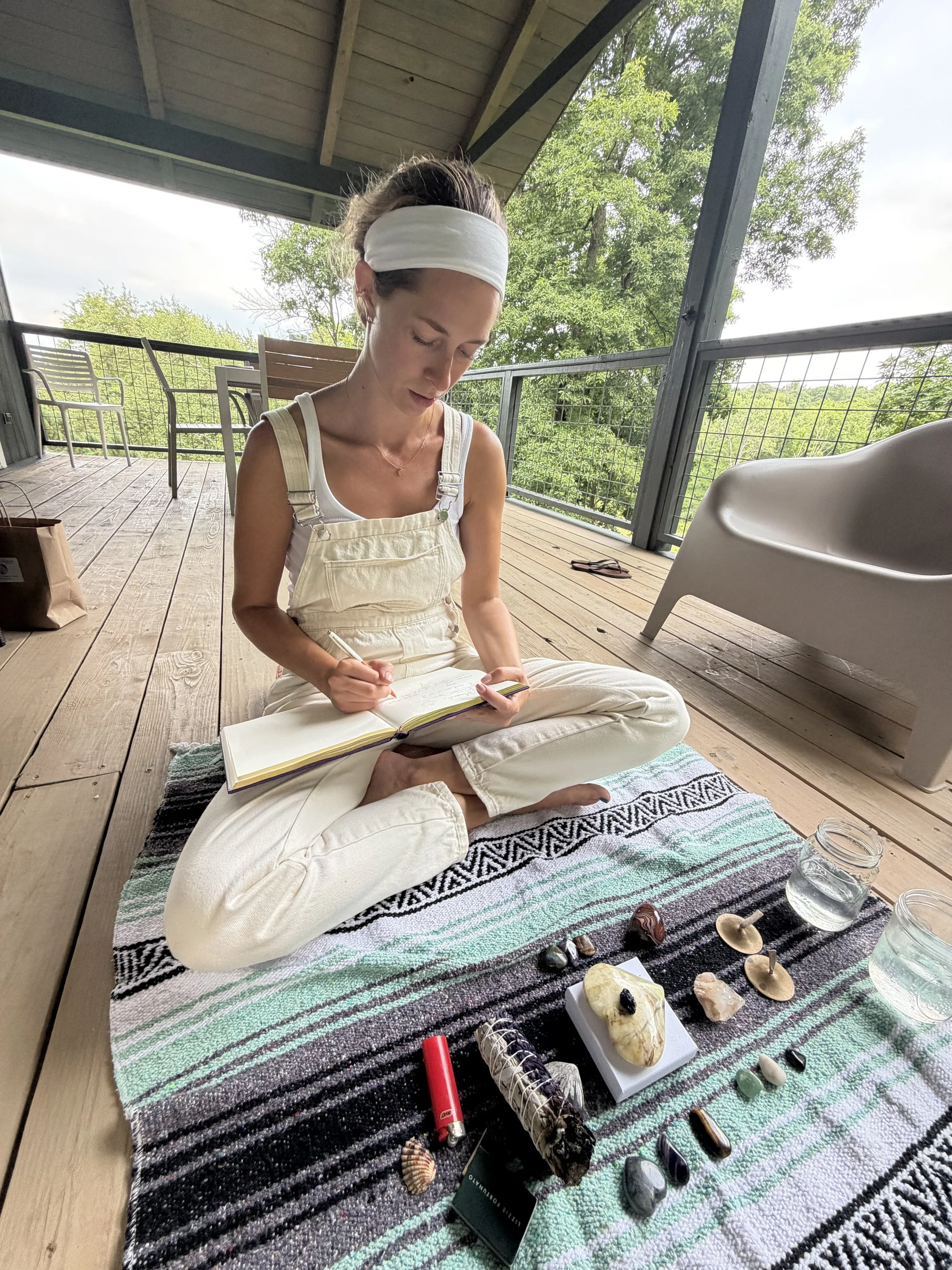 A woman with a white headband and white overalls sitting cross-legged on a striped blanket on a wooden porch, writing in a notebook with various crystals, stones, and decorative items arranged in front of her.