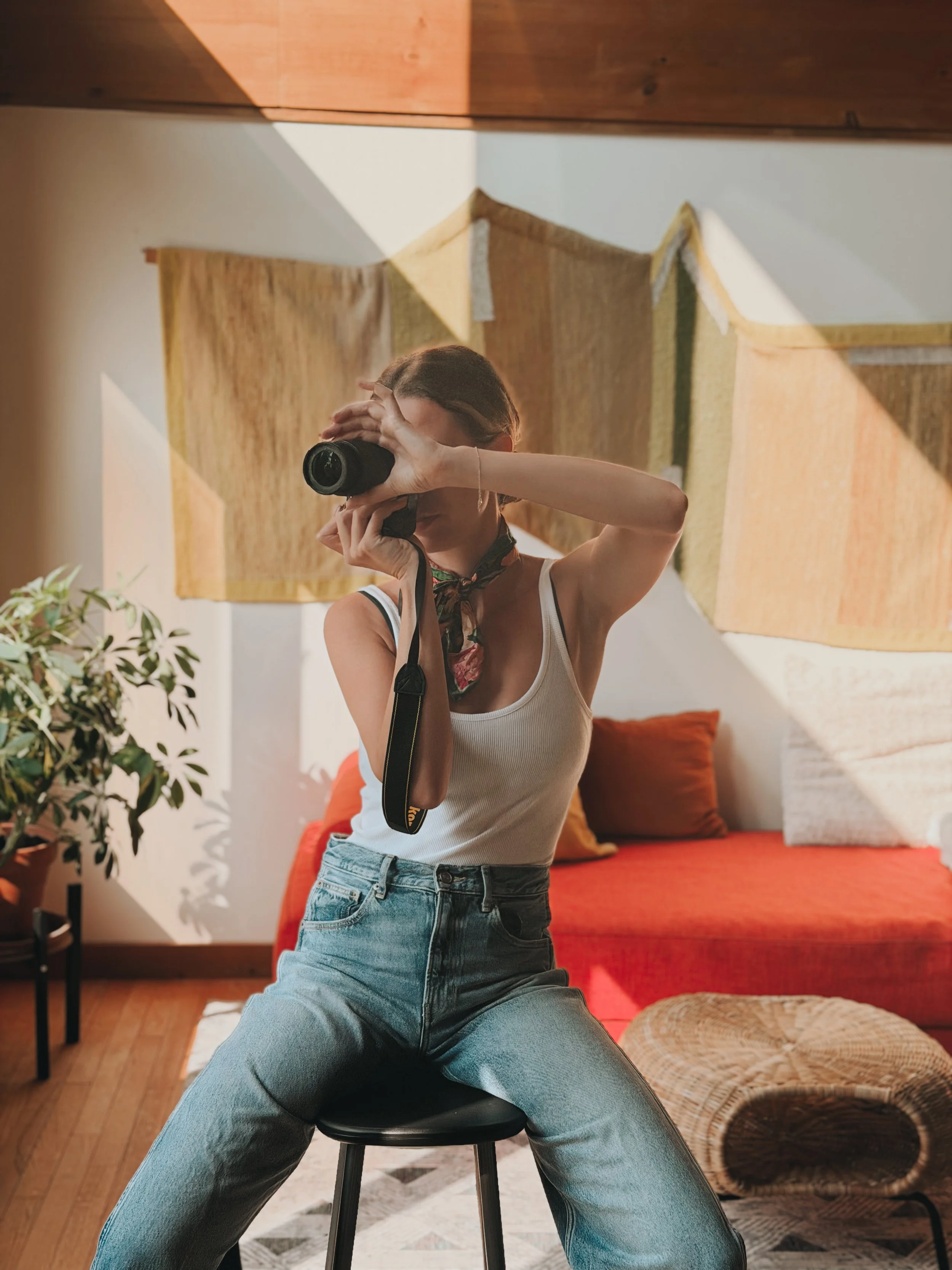 A person taking a photo while seated on a stool, wearing a white tank top, jeans, and a scarf around the neck, with a living room background and decorative wall hangings.