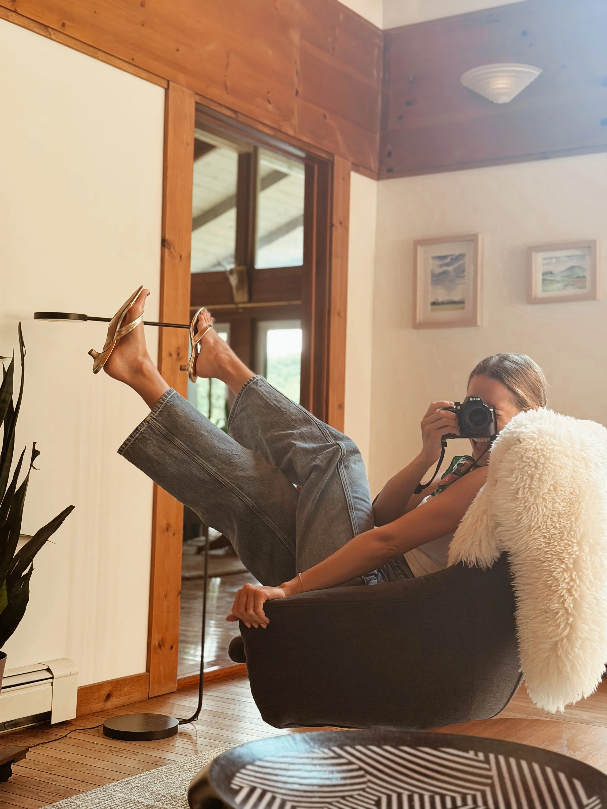 Person lying on a chair with feet up against the wall, wearing sandals, with a person taking a photo of their feet using a camera. The scene is in a cozy room with wooden accents and framed pictures on the wall.