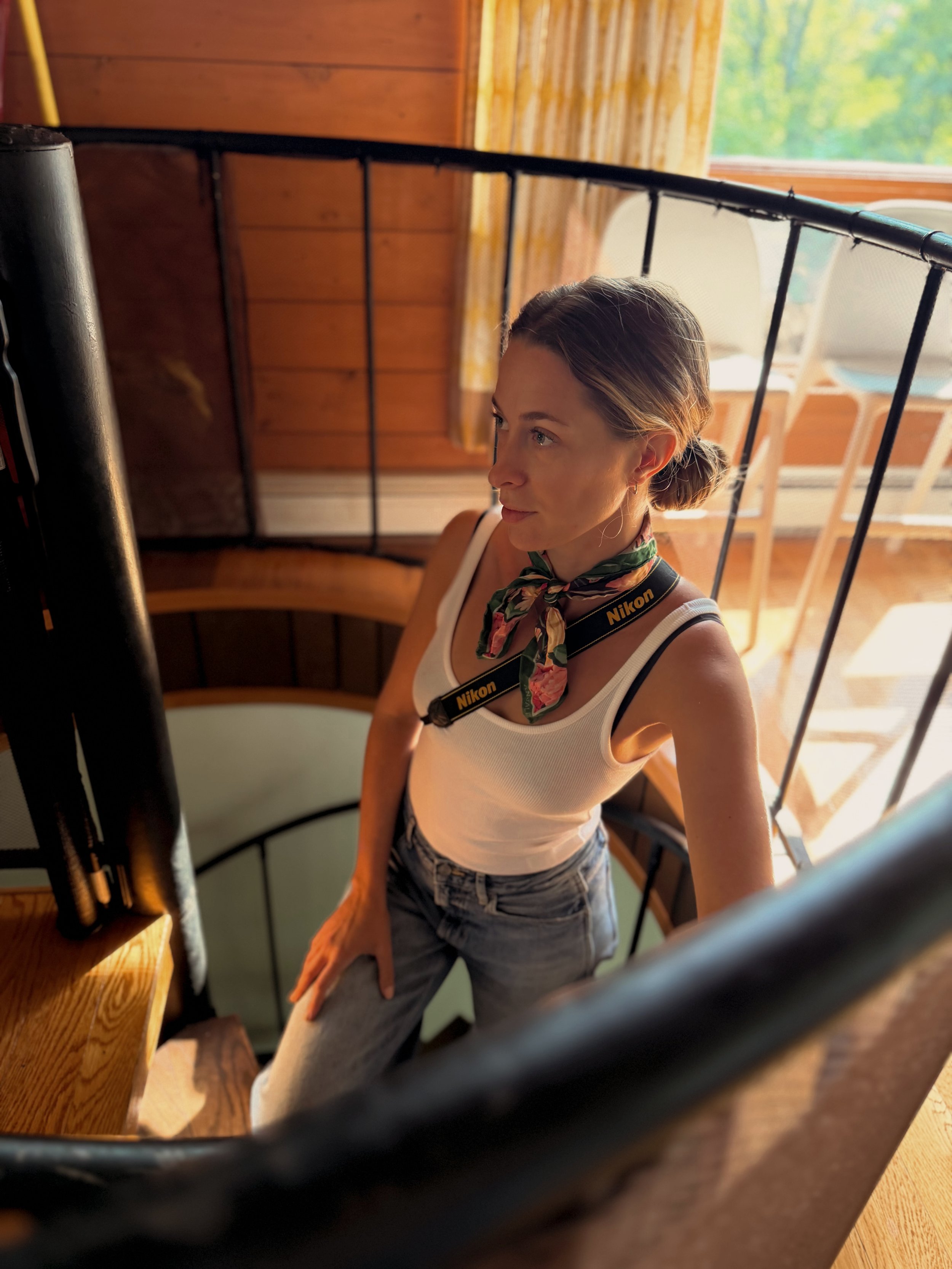 A woman with brown hair pulled back, wearing a white tank top and denim shorts, is looking out a window through a mirror. She has a Nikon camera strap around her neck and a colorful scarf tied around her neck. The room has wooden walls and a table with chairs outside.