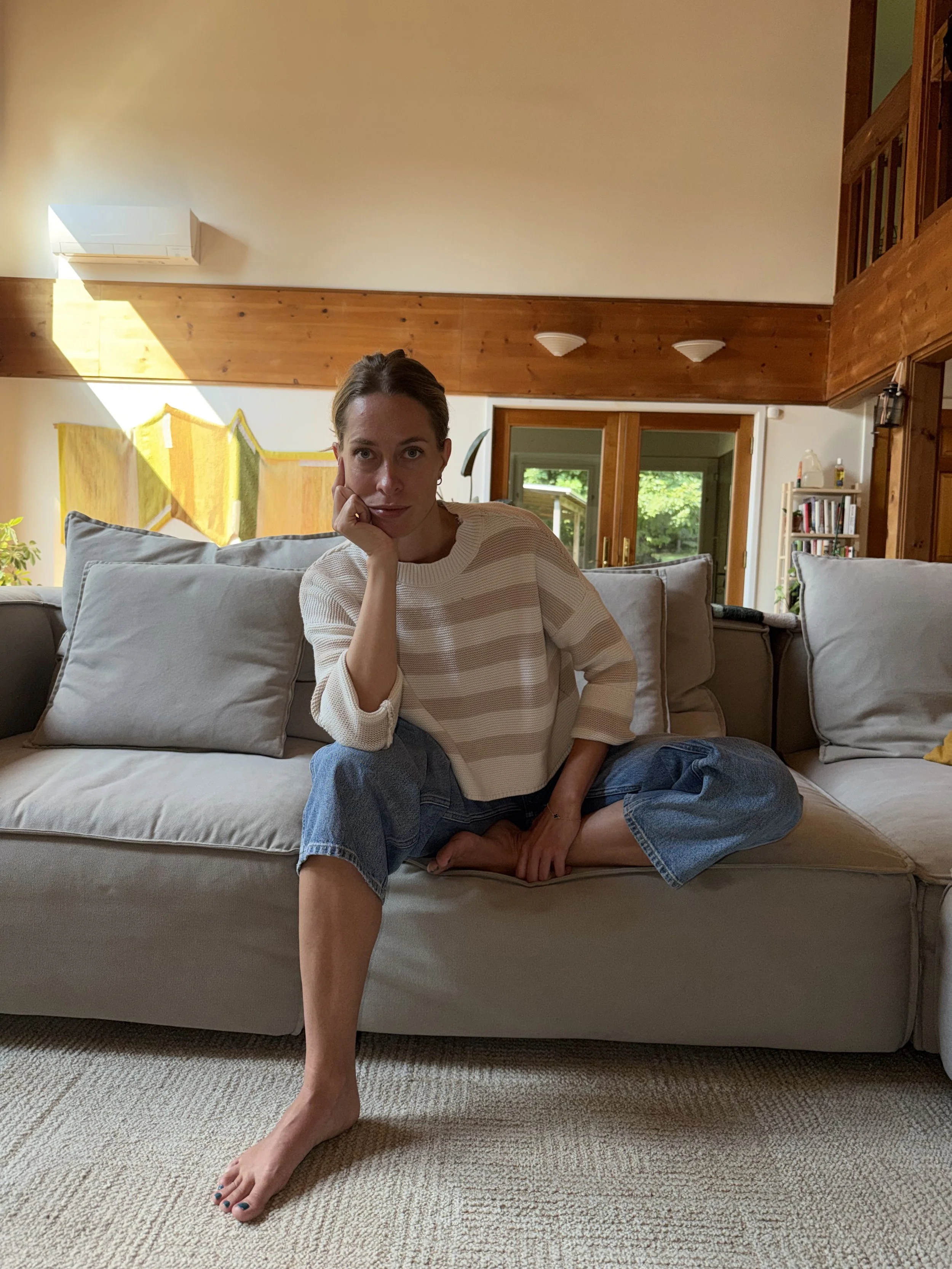 A woman sitting on a couch in a living room with her right hand resting on her face, wearing a beige striped sweater and jeans, with a multi-story house visible through large windows behind her.