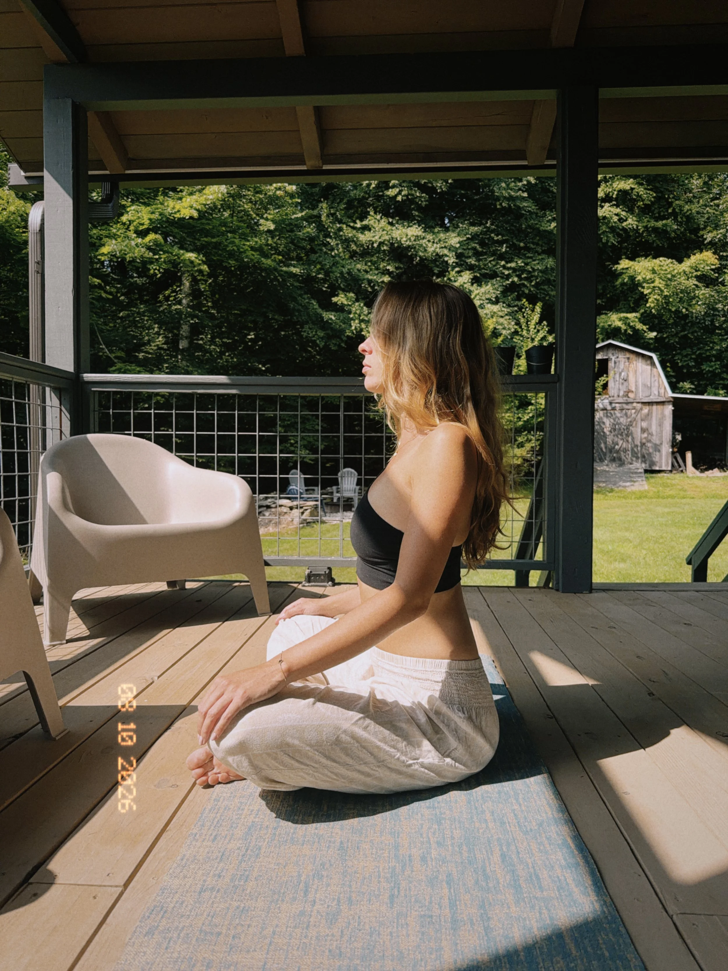 A woman with long hair, wearing a black top and white pants, is sitting cross-legged on a yoga mat outdoors on a wooden deck, meditating with her eyes closed.