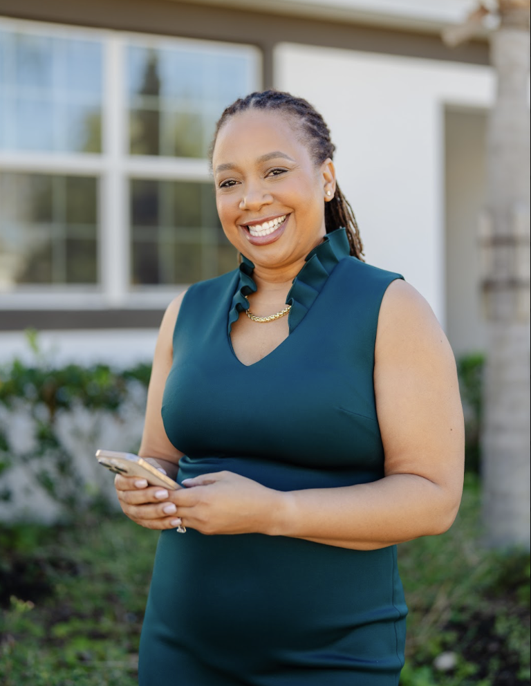 A smiling woman with braided hair wearing a sleeveless teal dress holding a phone outdoors in front of a house with large windows and greenery.