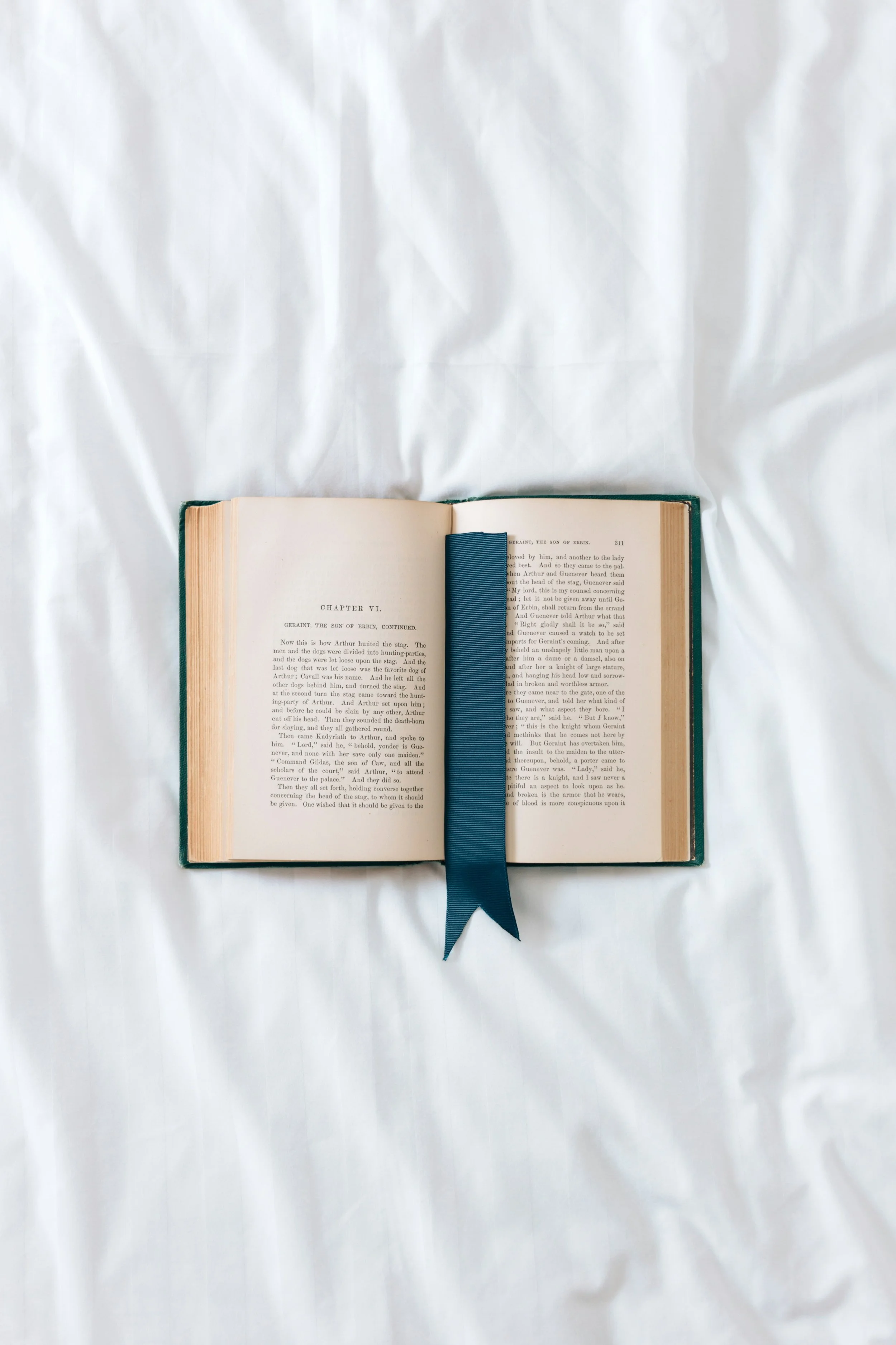 An open hardcover book with aged pages, a blue bookmark ribbon, and a green cover, placed on a white wrinkled fabric background.