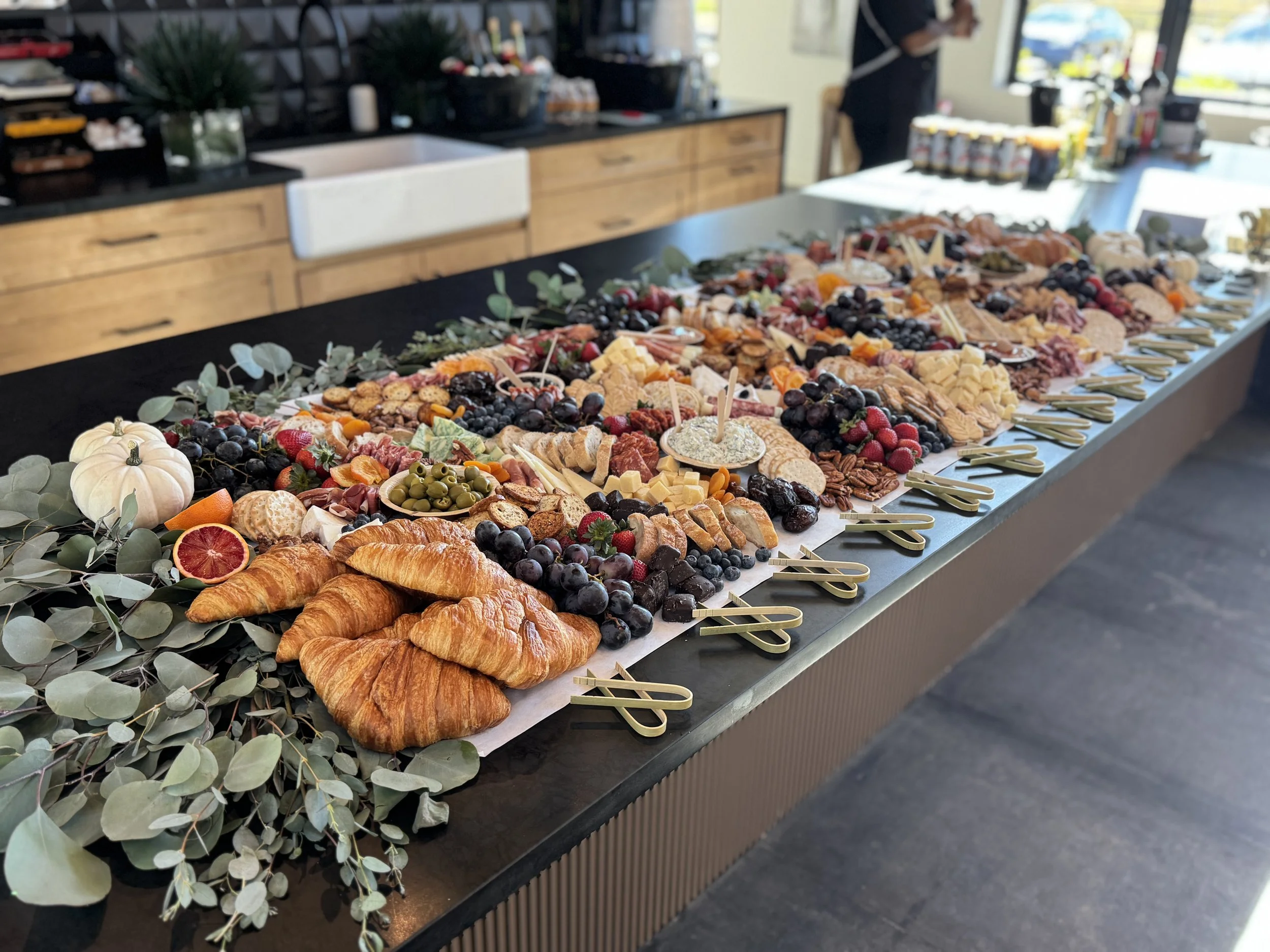A buffet table set with various cheeses, grapes, berries, crackers, and pastries, decorated with eucalyptus leaves, in a bright indoor setting.