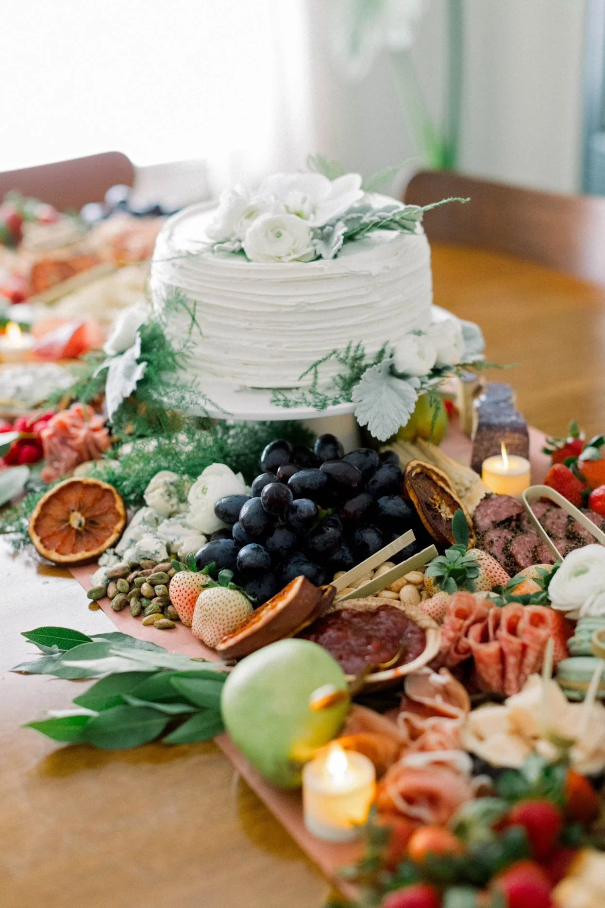 A white wedding cake decorated with white flowers, surrounded by assorted fruits and snacks on a long table, with candles and greenery for decoration.