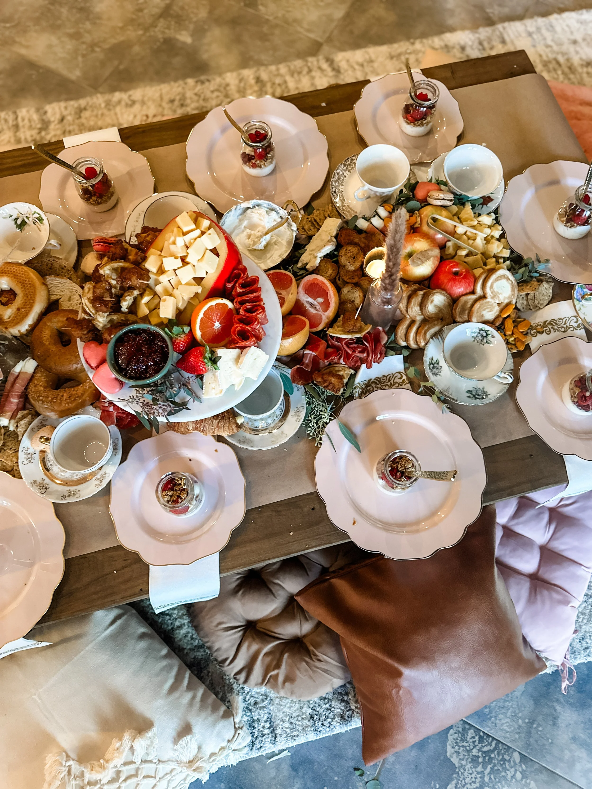 A breakfast table with assorted cheeses, sliced oranges, grapes, bread, pastries, jams, and bowls of yogurt, surrounded by empty teacups and glasses of water.