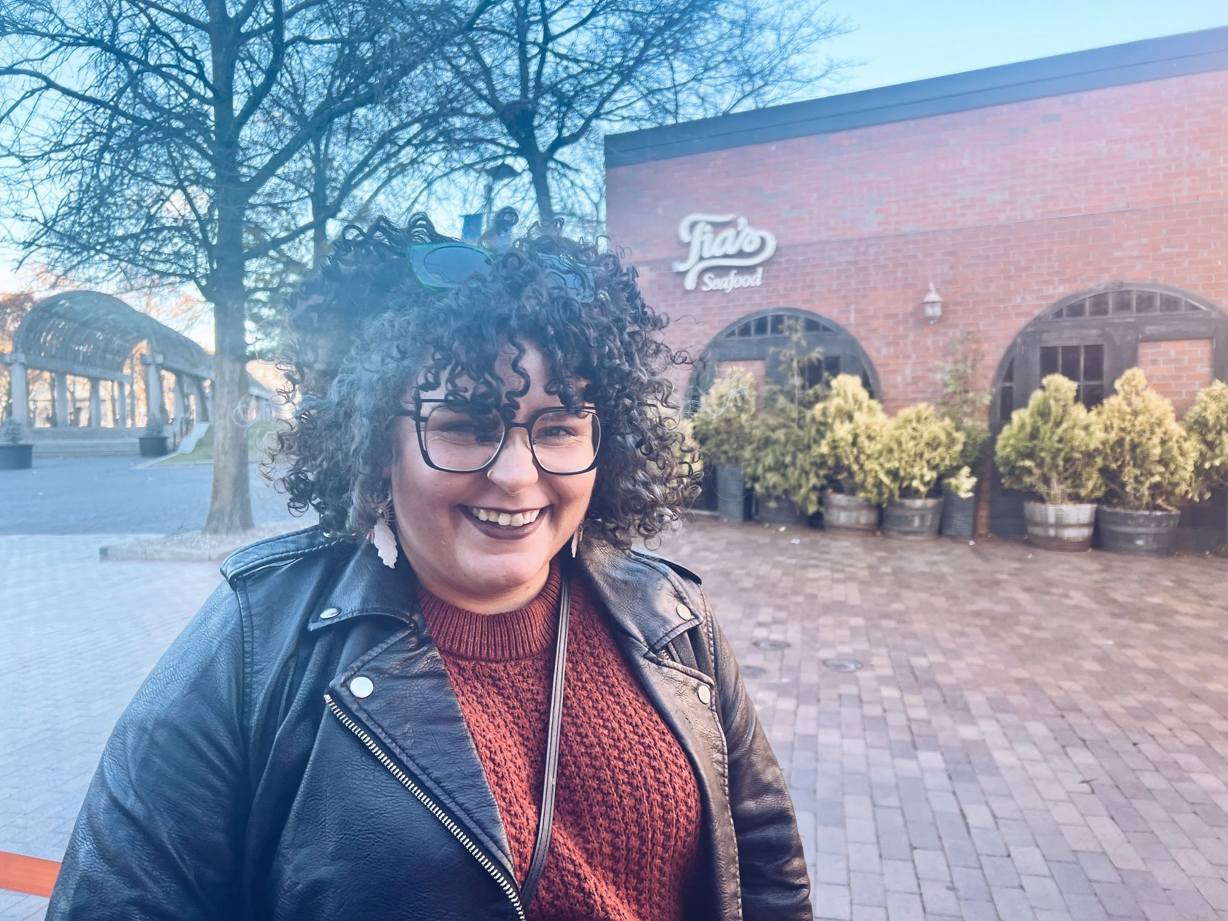 Smiling woman with curly hair, glasses, wearing a black leather jacket and a maroon sweater, standing outdoors in front of a brick building with a sign that says 'Tiamo Seafood' and trees.