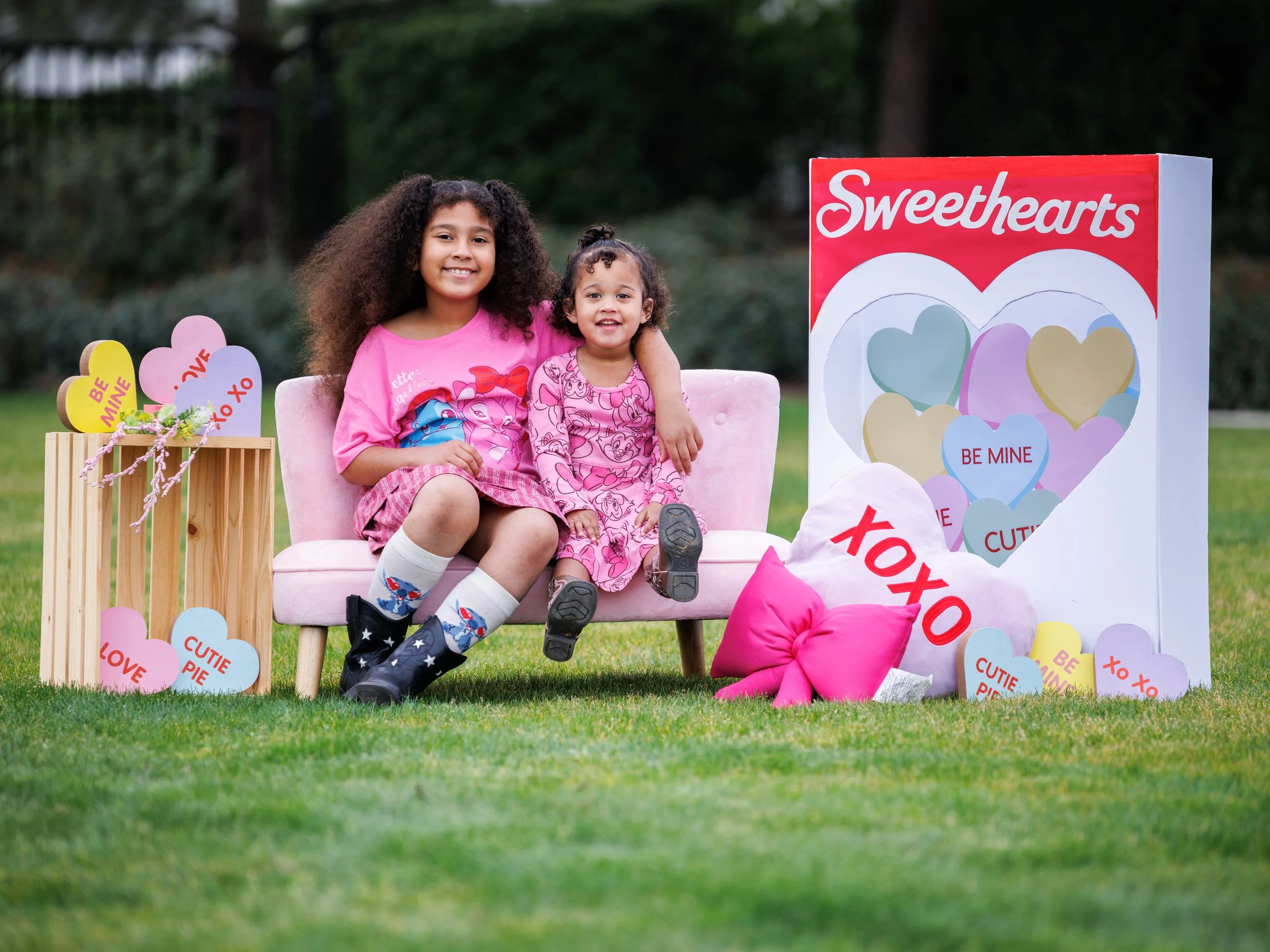 Two girls sitting on a pink couch outdoors surrounded by valentine's day themed decorations, including hearts with words like 'LOVE' and 'BE MINE.' The older girl has curly hair and is wearing a pink shirt, while the younger girl has shorter curly hair and is also dressed in pink. There are colorful cushions and a small wooden table with more heart decorations.