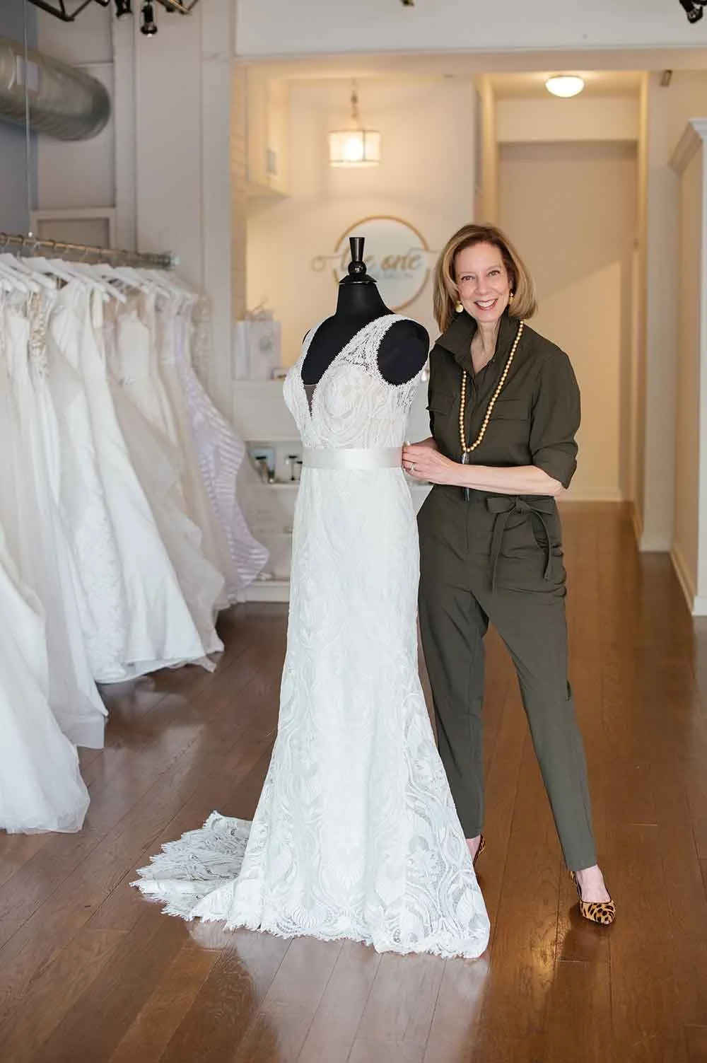 A woman standing next to a mannequin dressed in a white wedding gown inside a bridal shop. The woman is smiling and wearing an olive green jumpsuit with leopard print heels, a gold necklace, and earrings. Bridal dresses are hanging on a rack on the left side of the image.