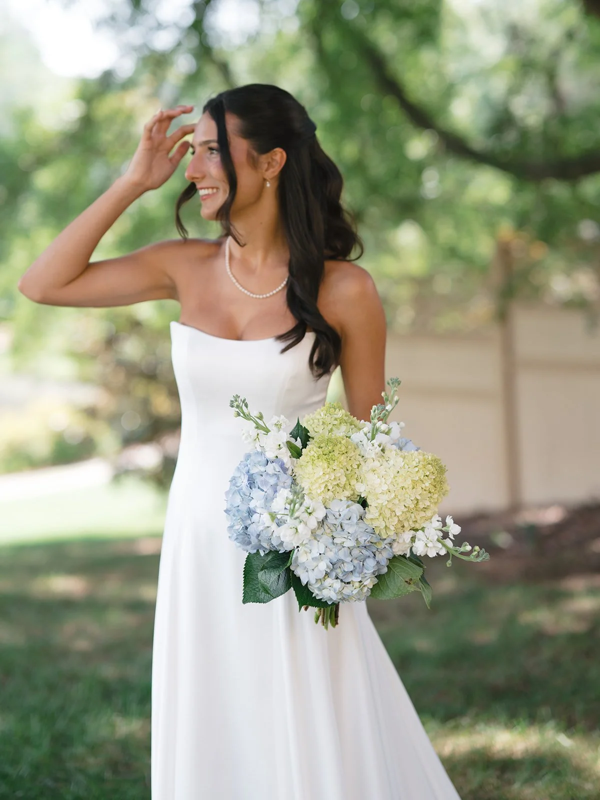 A woman in a white strapless dress holding a bouquet of hydrangeas, smiling outdoors with trees in the background.