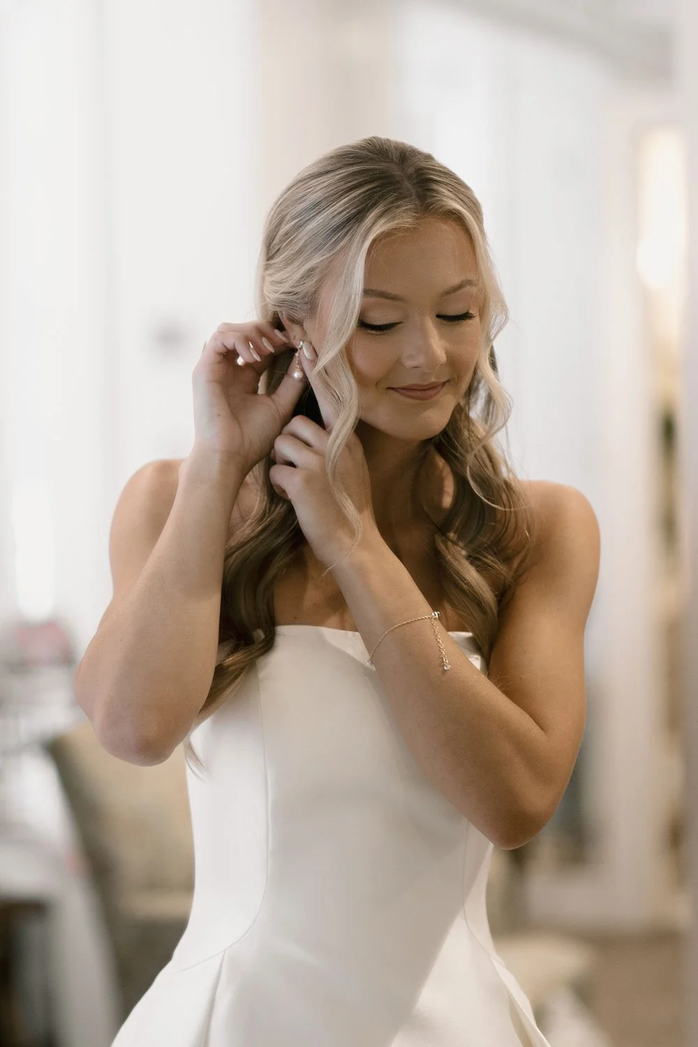 Bride adjusting her earring, wearing a white wedding dress, with long wavy hair, smiling softly.