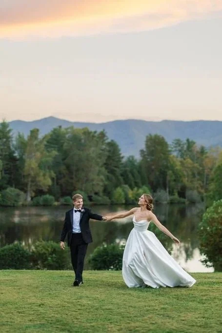A bride and groom dancing outdoors near a lake with trees and mountains in the background during sunset.