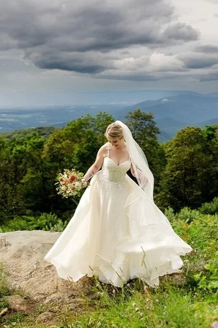 Bride in a white wedding gown holding a bouquet, standing outdoors on a rock with a mountain and cloudy sky background.