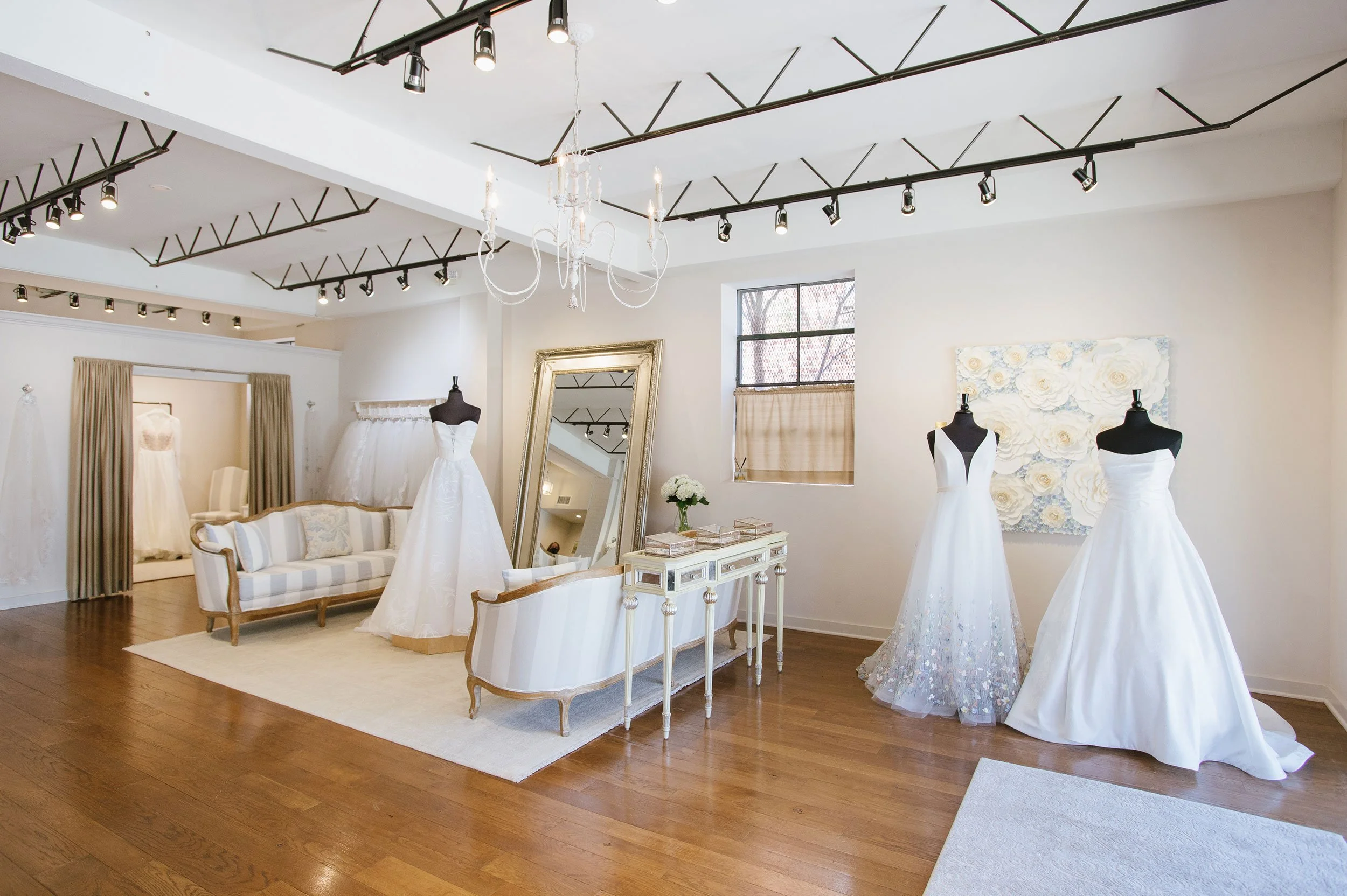 A bridal boutique with wedding dresses on mannequins, a large mirror, a chandelier, and a white and beige color scheme.