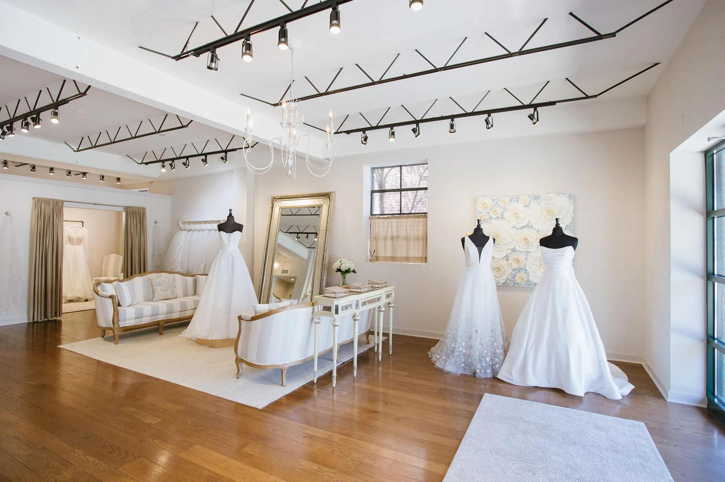 A bridal boutique with wedding dresses on mannequins, a large ornate mirror, a vintage sofa, and a chandelier, with bright natural light coming through the windows.