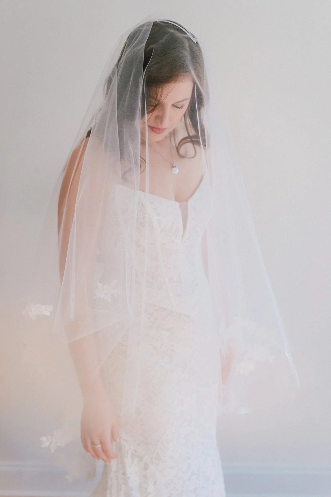 A woman in a white lace wedding dress with a long veil looking down, standing against a plain white wall.