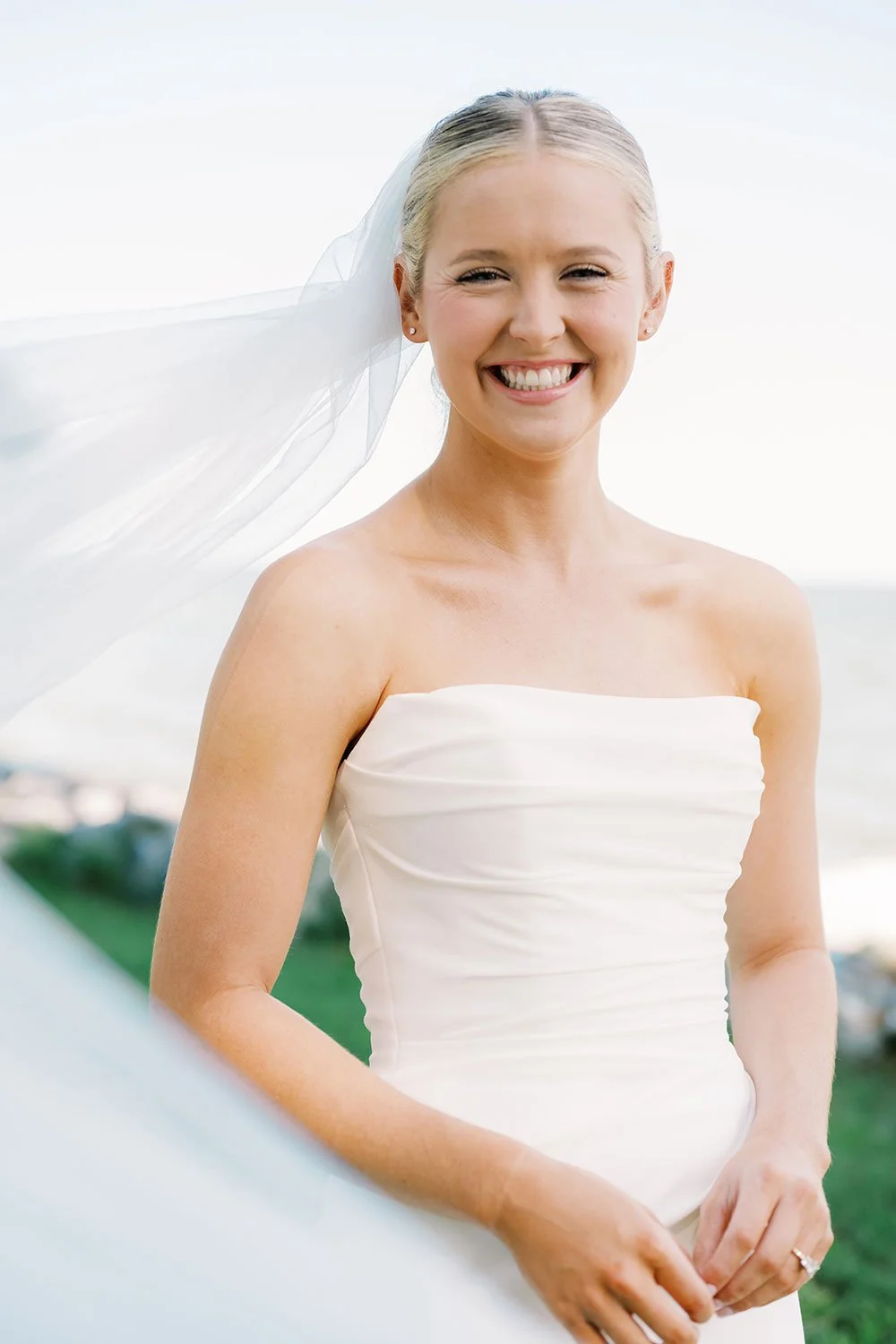 A smiling bride in a white strapless wedding dress outdoors with a veil flowing in the breeze.