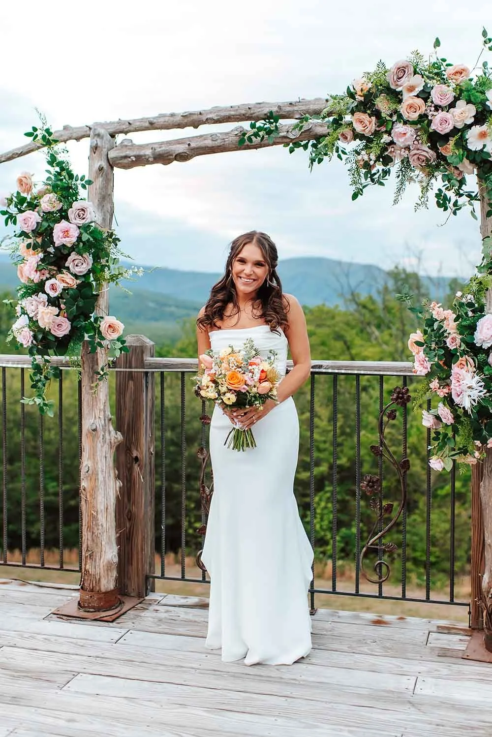 A young woman in a white wedding dress holding a bouquet of flowers, standing under a floral arch on a wooden platform outdoors with mountains and cloudy sky in the background.