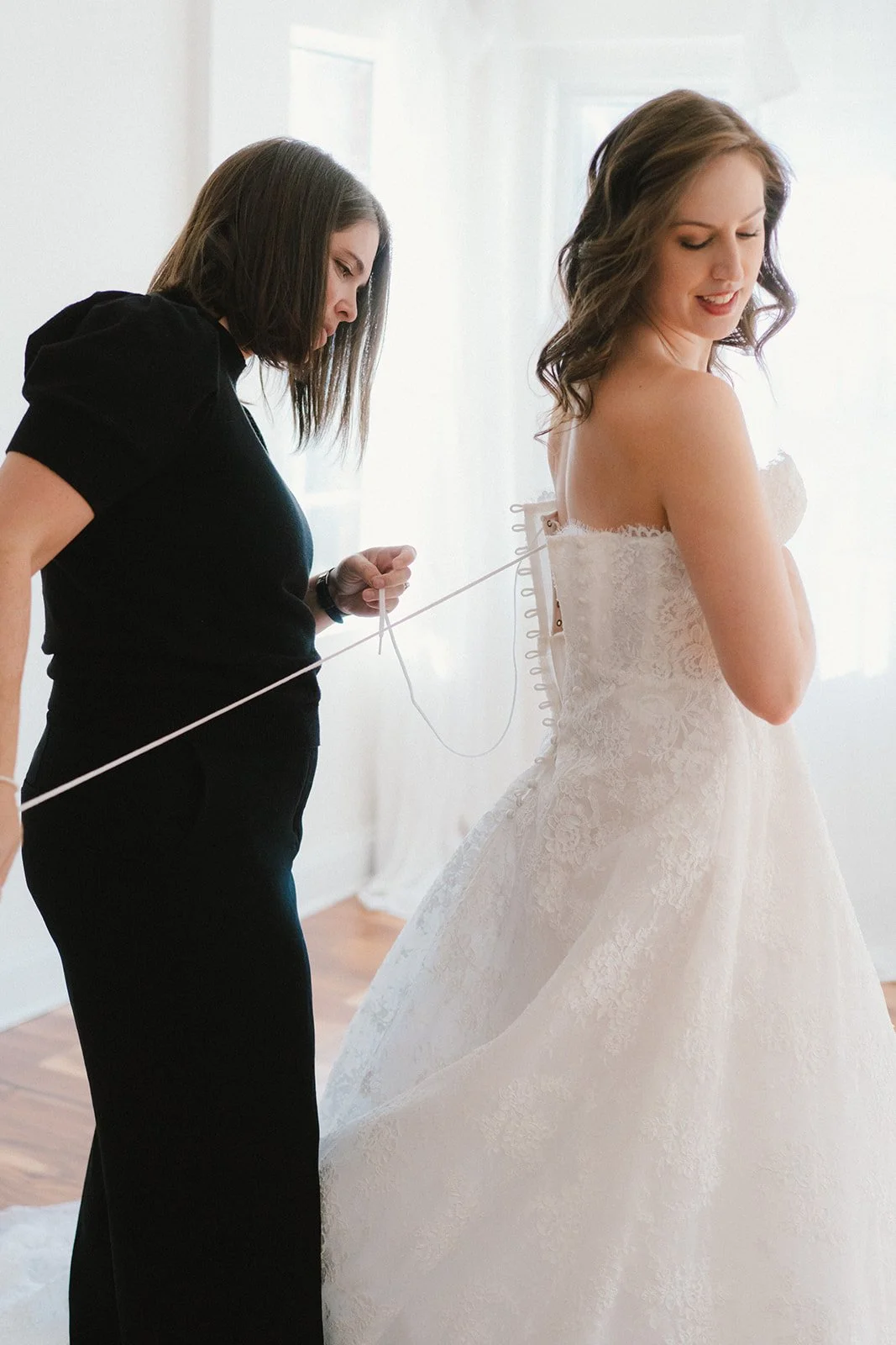A bride in a white lace wedding dress having her zipper fastened by a woman in black, standing in a bright room.
