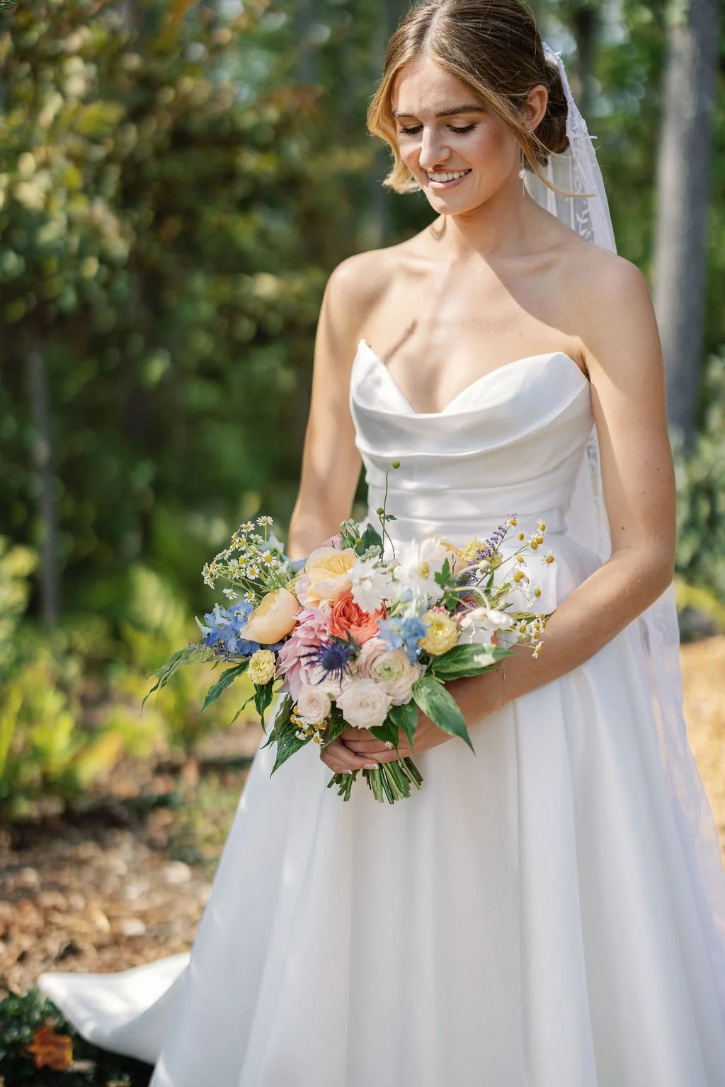 A smiling bride in a white wedding gown holding a colorful bouquet of flowers outdoors.