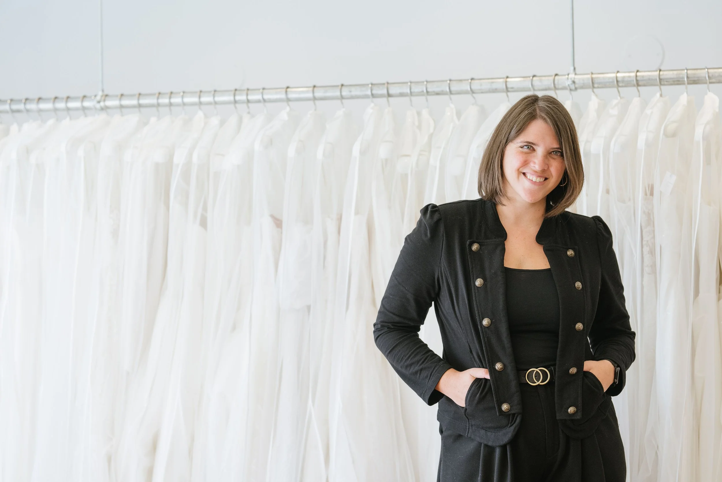 A woman with shoulder-length brown hair smiling while standing in front of a rack of white dresses or gowns.